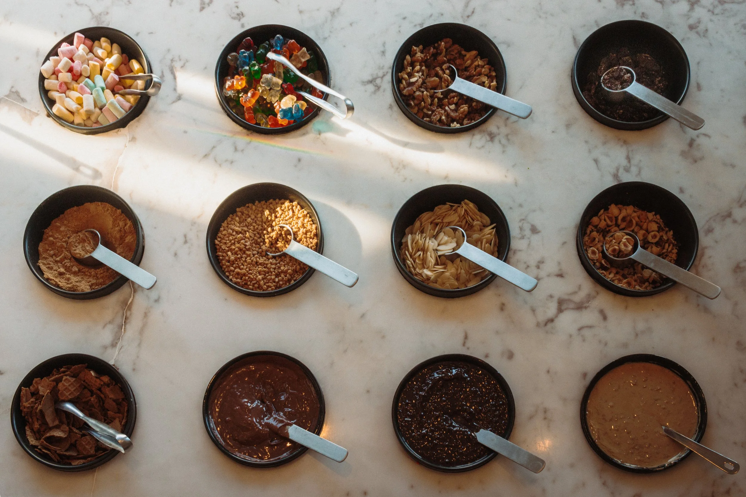 Assorted toppings in small black bowls on a white marble surface, including marshmallows, gummy bears, chopped nuts, chocolate powder, crushed cookies, caramel bits, and various sauces.