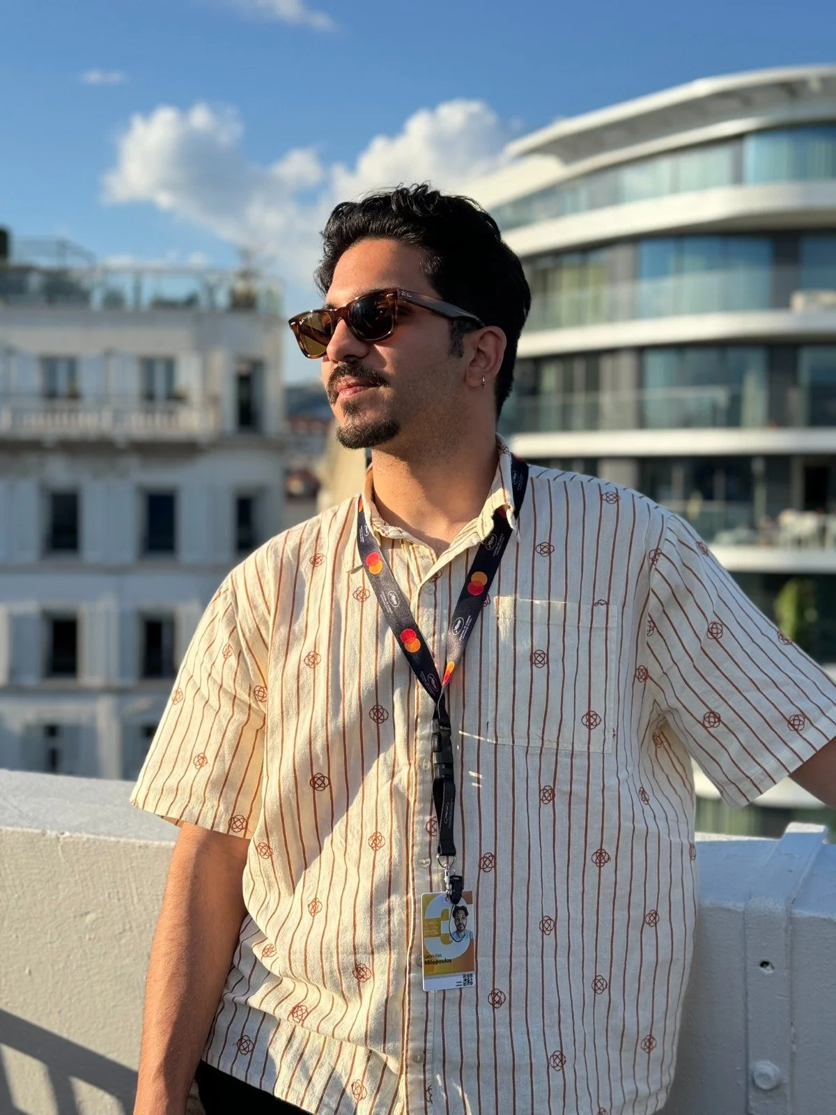 A man with dark hair and a mustache wearing sunglasses, a striped shirt, and a pass hanging from a lanyard, standing on a rooftop with city buildings in the background on a sunny day.