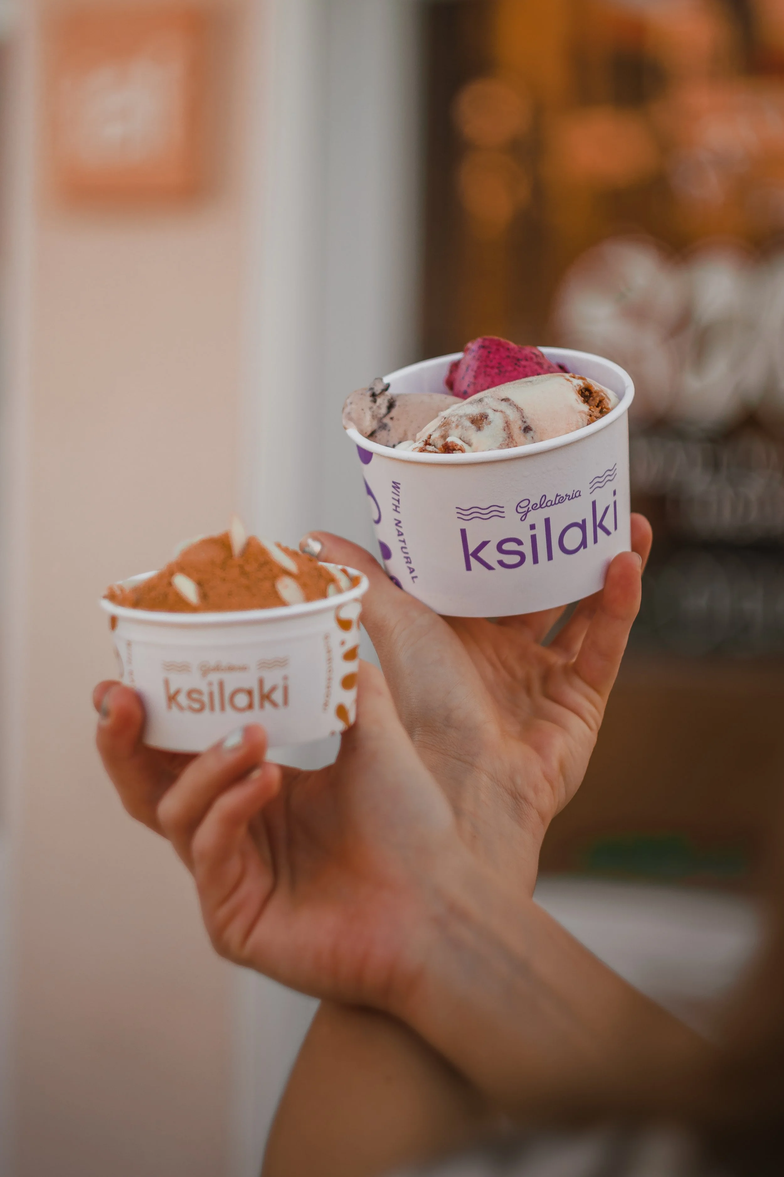 Two hands holding cups of gelato with different flavors at a gelateria named 'Ksilaki', with one cup in focus showing a scoop of pink, beige, and chestnut-flavored gelato.