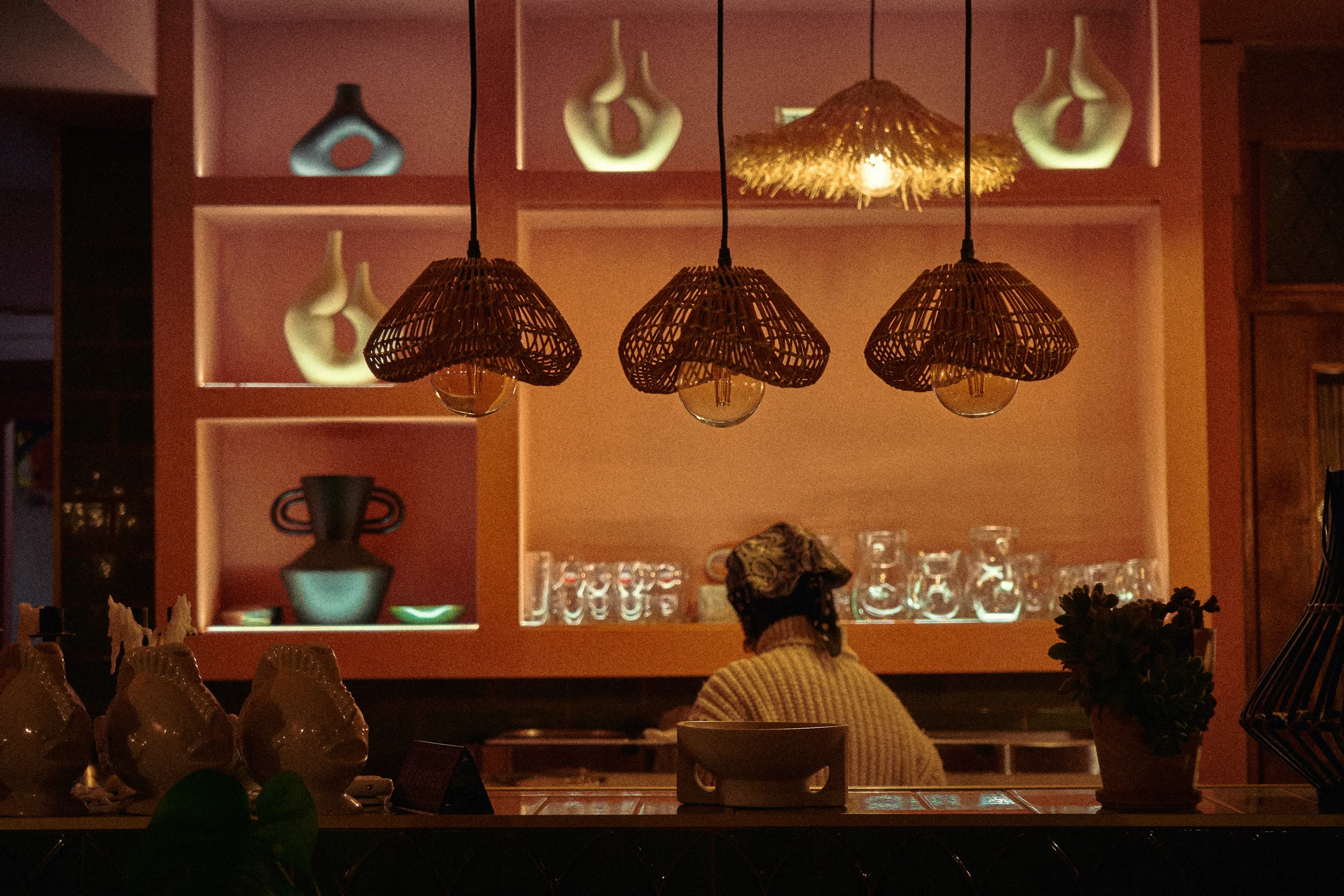 Indoor scene with hanging wicker pendant lights above a bar or countertop. Shelves in the background display decorative vases, glassware, and dishes, with warm lighting creating a cozy atmosphere.