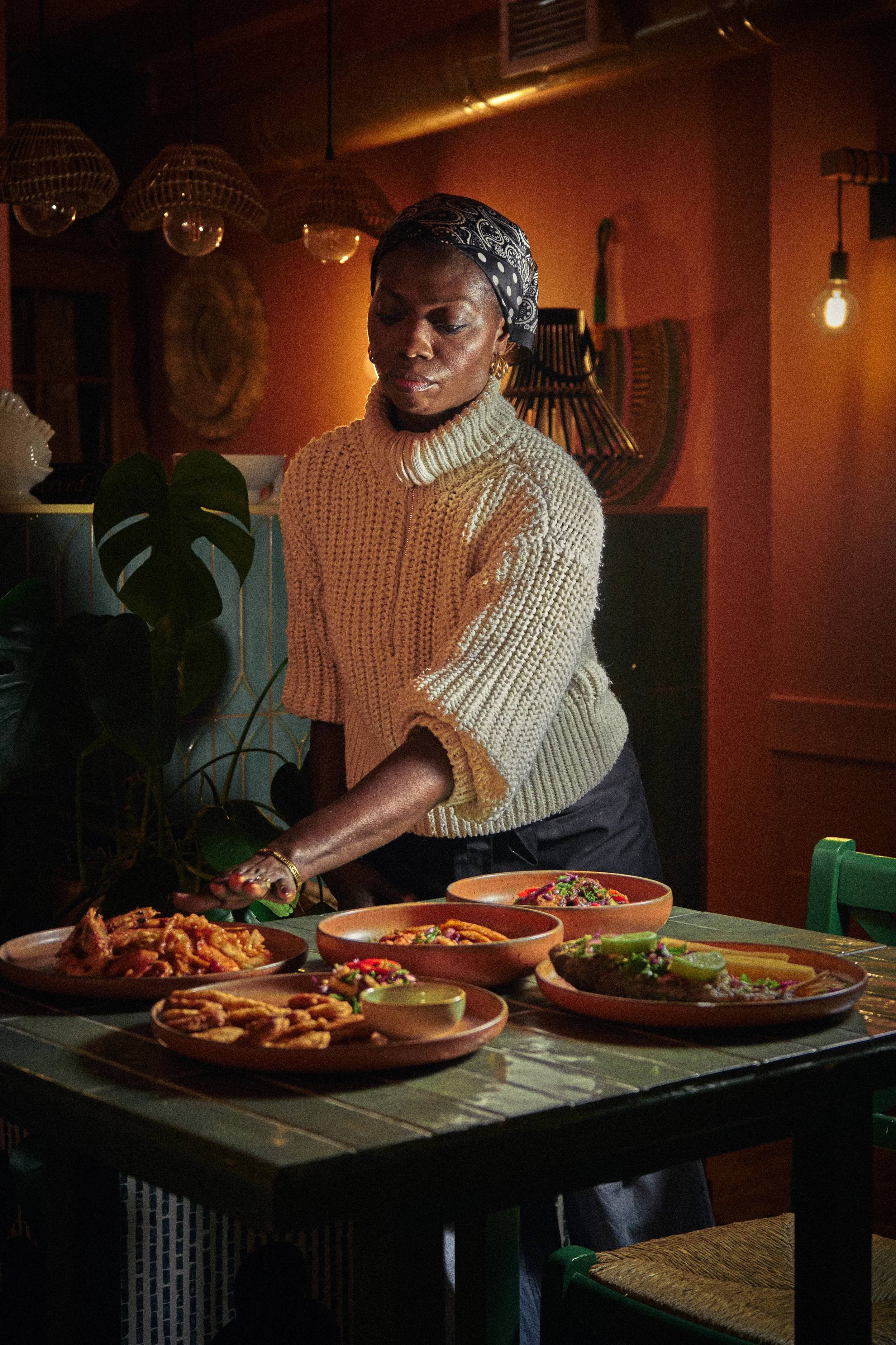 A woman with a headscarf and jewelry serving various dishes of food on a table, in a warmly lit room with decorative items and bonsai on the wall.