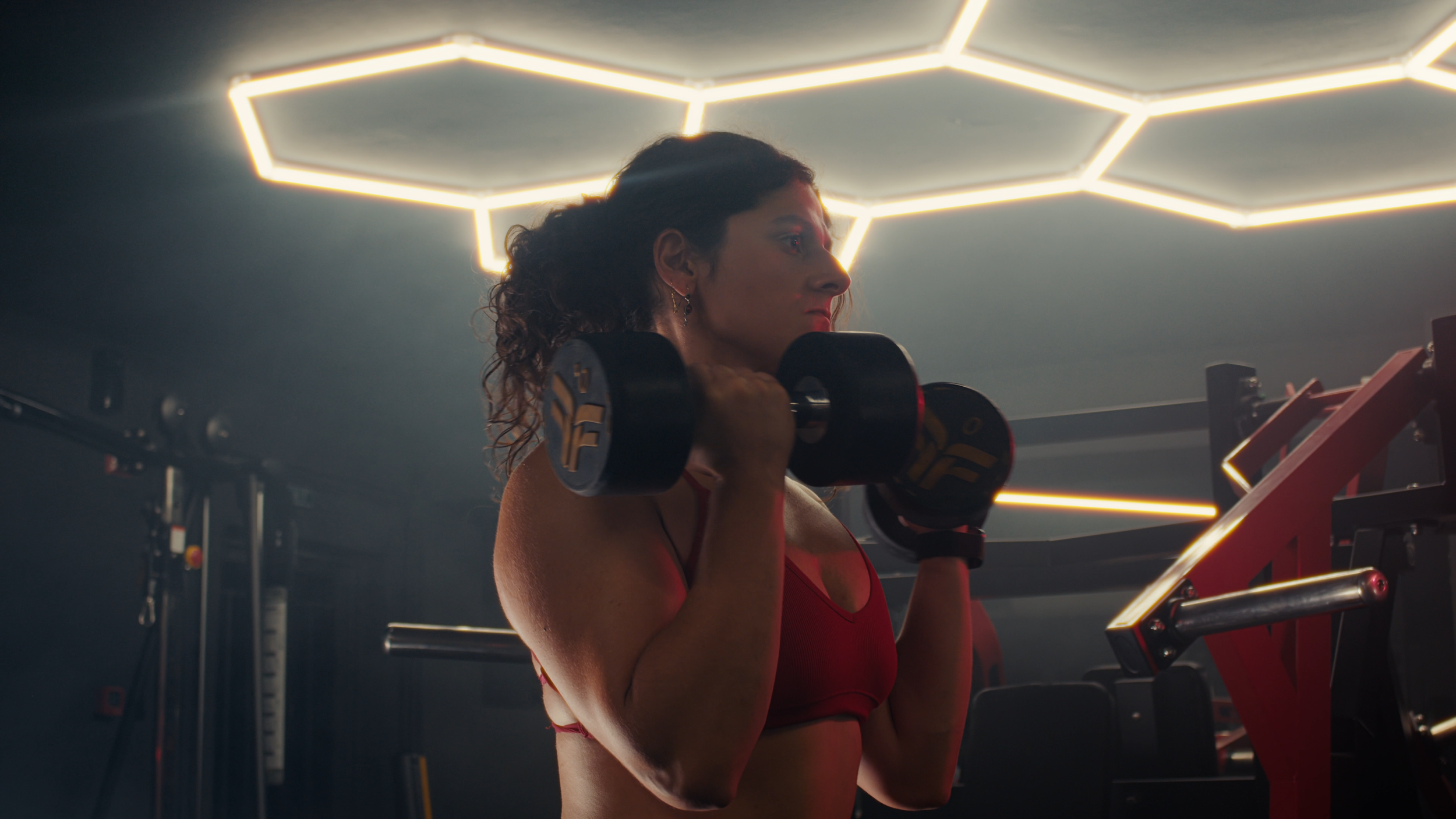 A woman with curly hair and earrings lifting a dumbbell in a gym with modern hexagonal lighting on the ceiling.