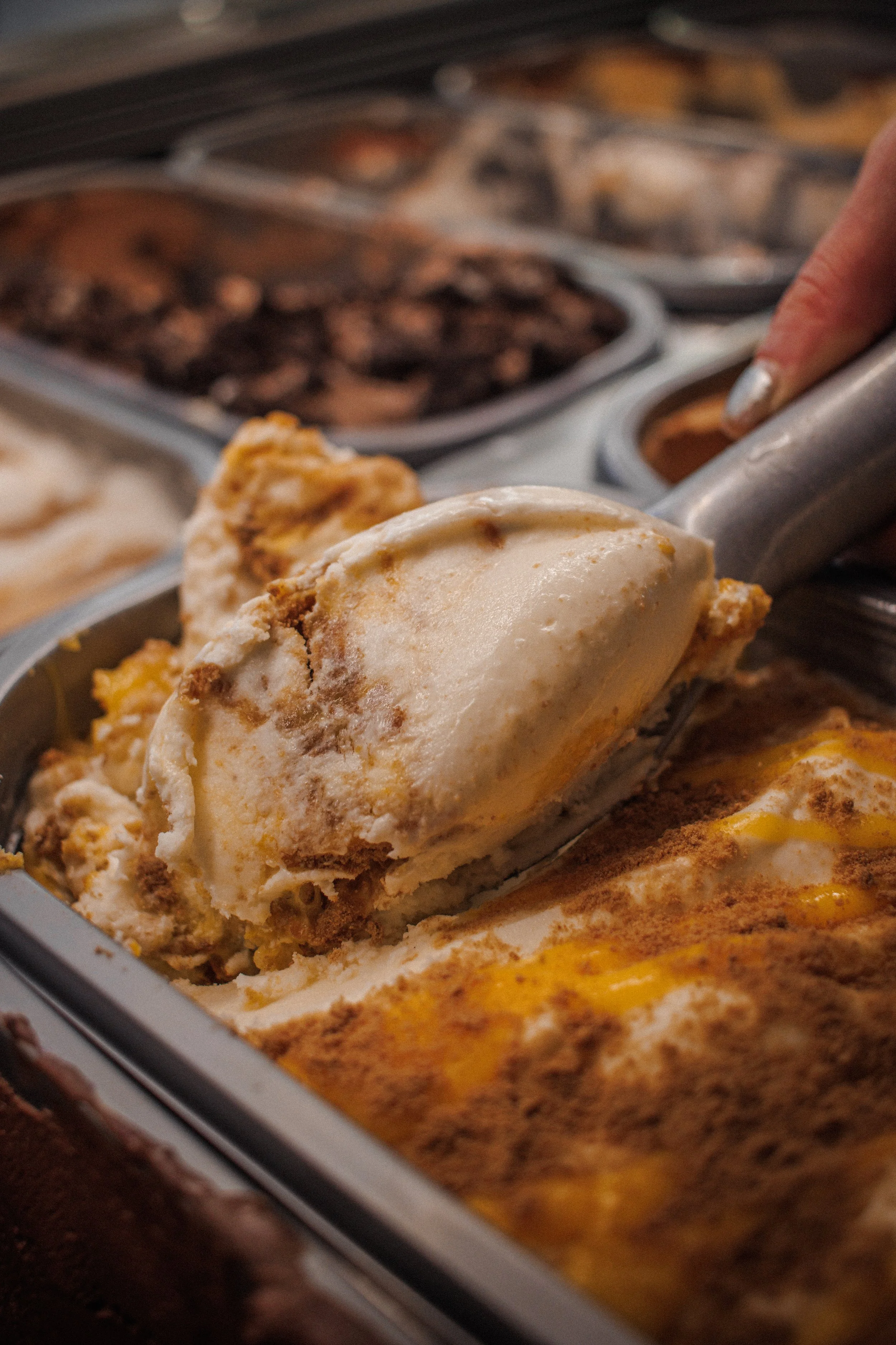 Ice cream scoop with cookies and cream flavor in front of a variety of ice creams in metal containers, with chocolate chips and caramel flavors visible.