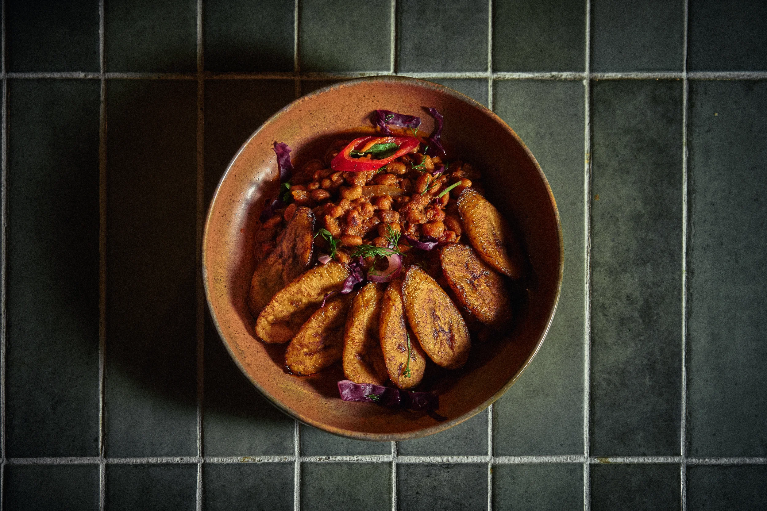 A bowl of cooked lentils with fried plantains, garnished with sliced red chili and green herbs, placed on a dark tiled surface.