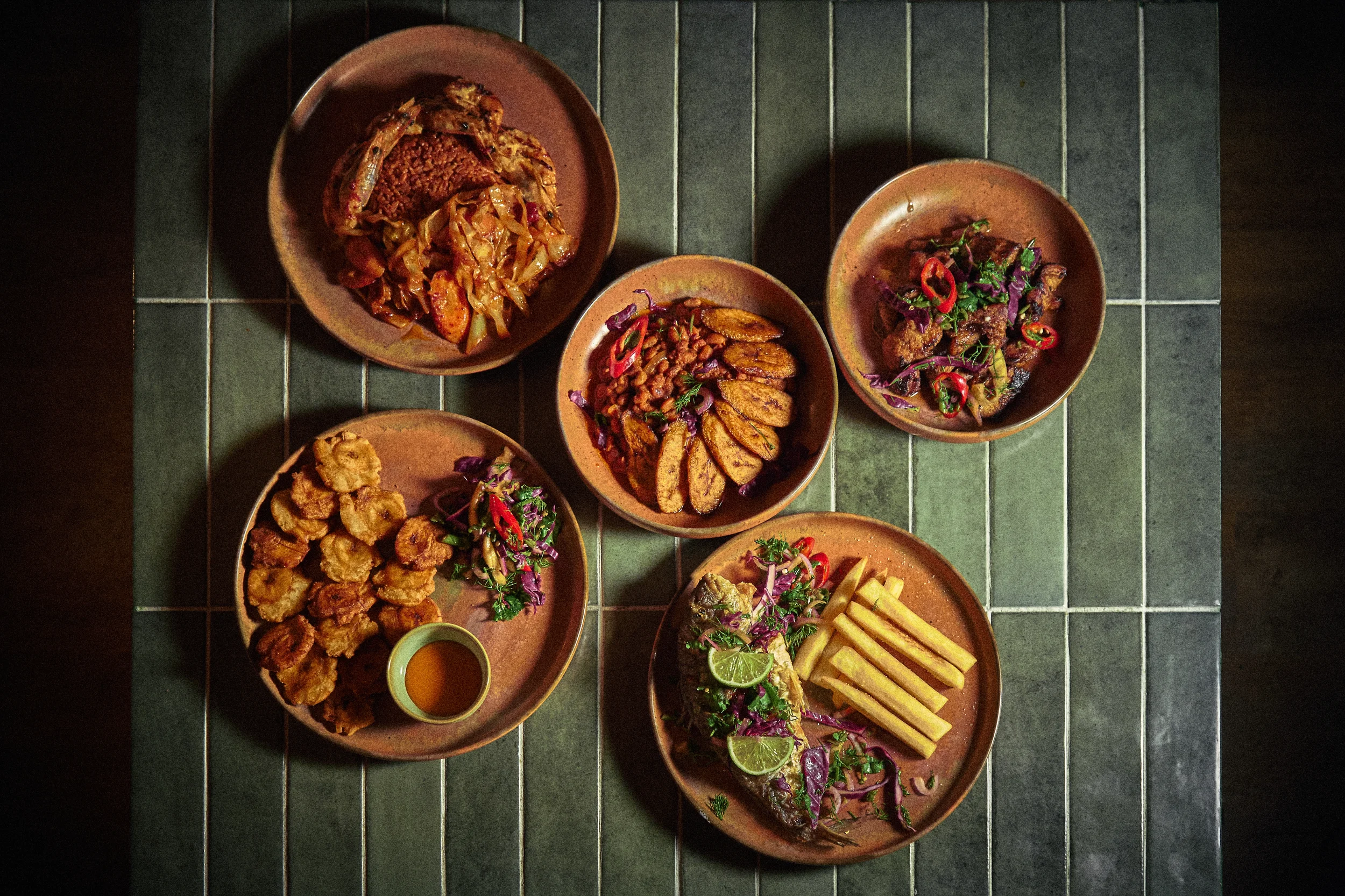 Assorted plates of Mexican food including fried chicken, potato wedges, fried fish, and other dishes with vegetables and lime wedges on a dark tiled table.