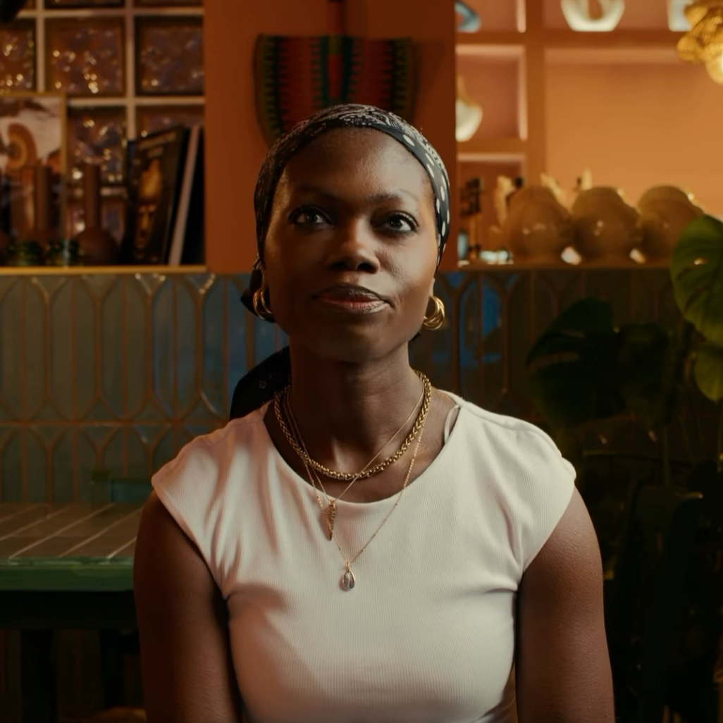 A woman with dark skin, wearing a white top, gold jewelry, and a patterned headscarf, sitting indoors with warm lighting and decorative items in the background.
