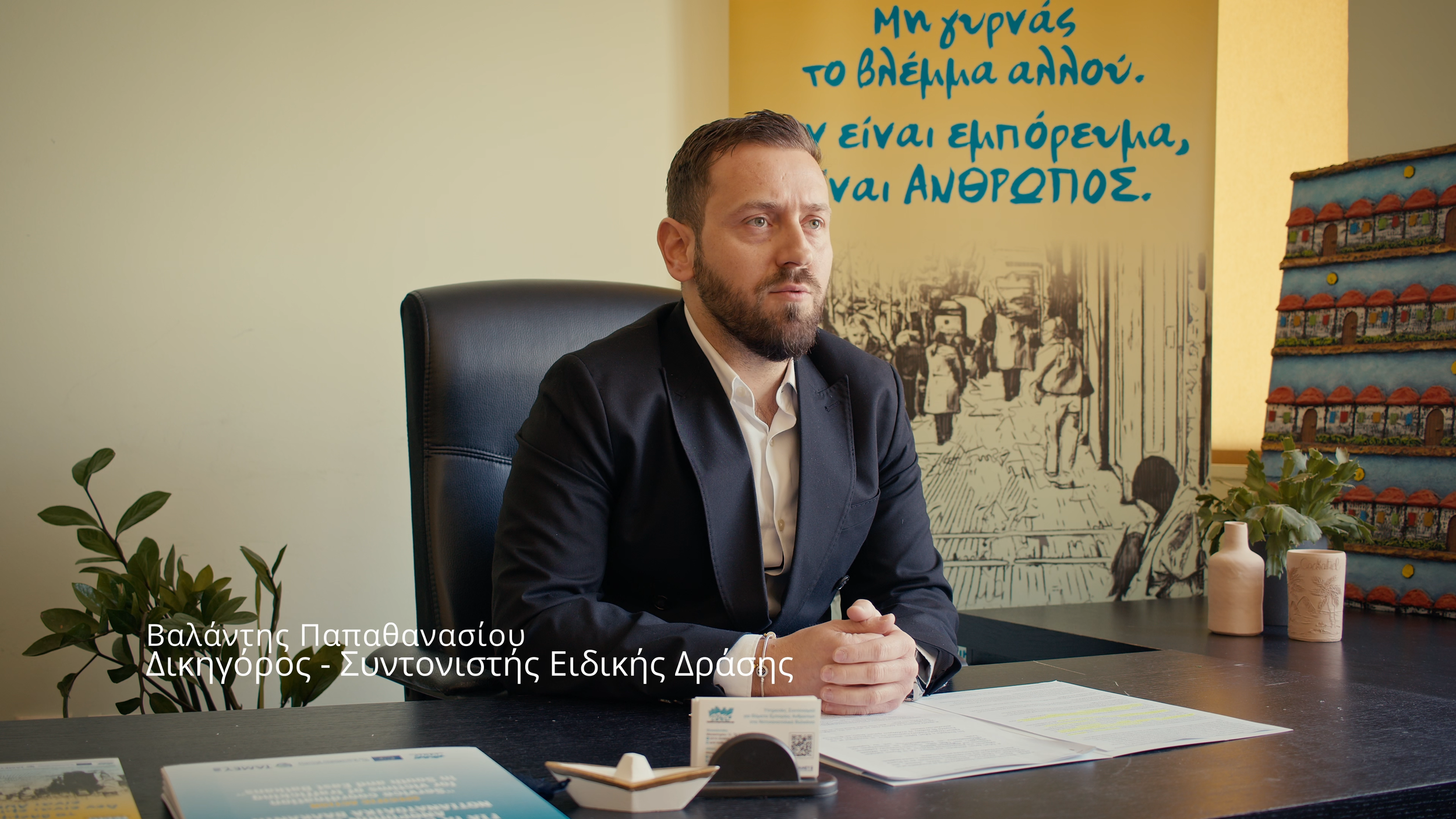A man in a dark suit and white shirt sitting at a desk, with a plant, papers, and small decorative items in front of him. Behind him is a yellow wall with blue Greek writing and a mural of people walking in a city. There are stacked colorful pottery pieces on the right.