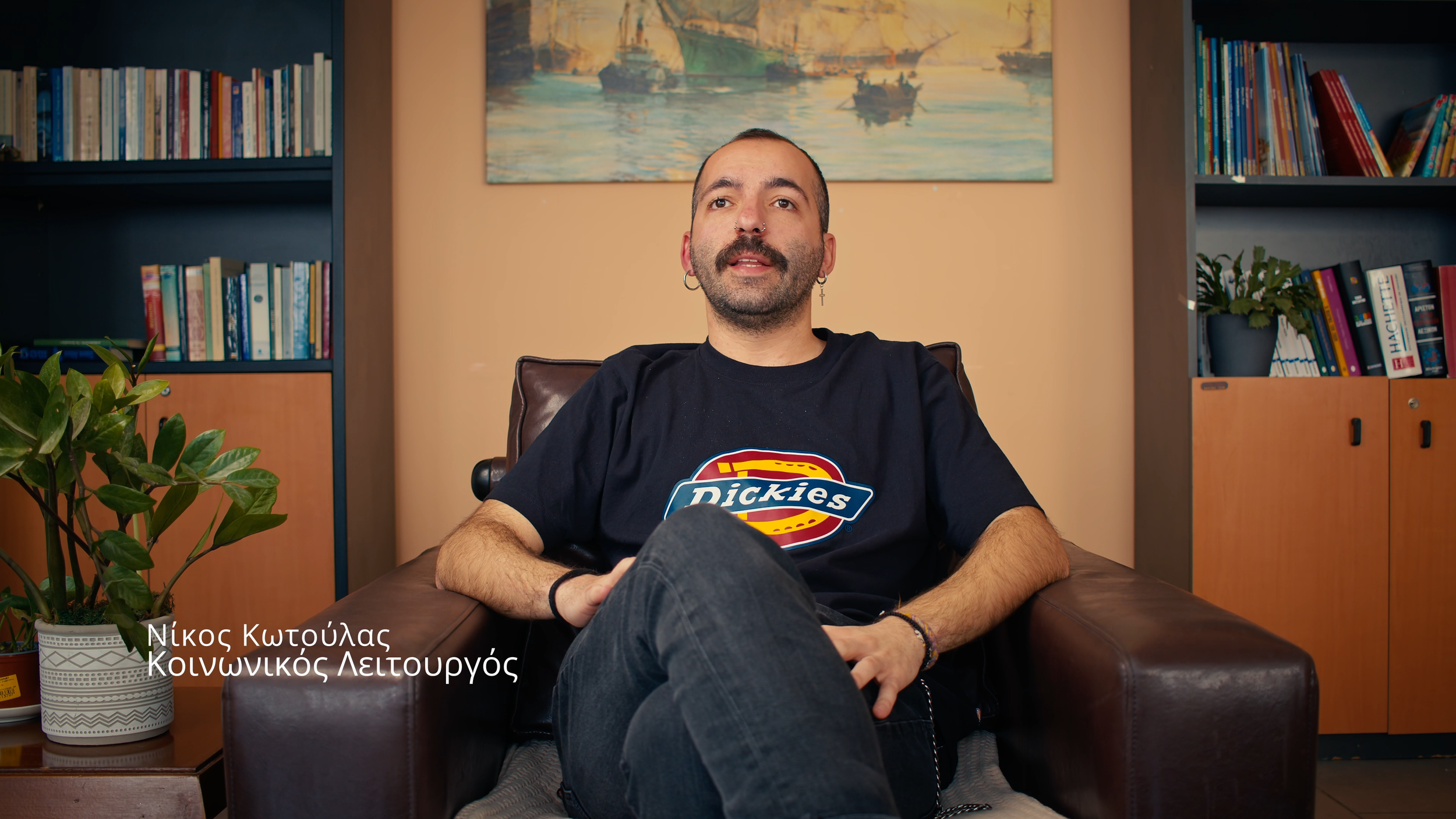 A man with a beard and earrings sitting in a brown leather armchair in a room with bookshelves, a potted plant, and a colorful landscape painting on the wall behind him. He is wearing a black Dickies t-shirt and has crossed legs.