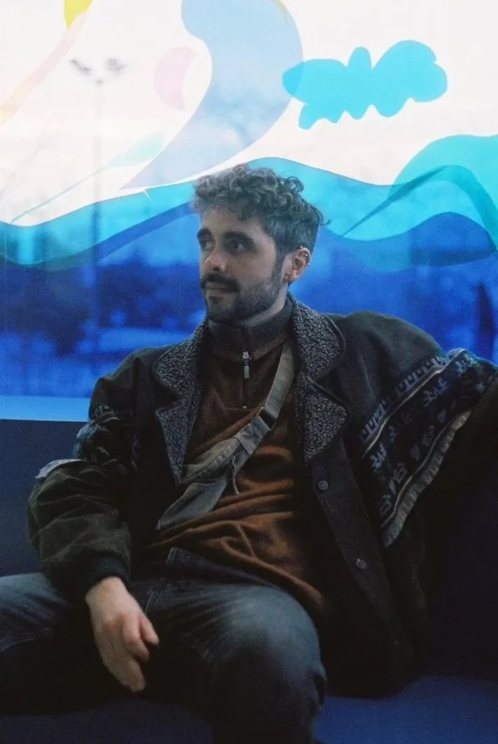 A man with curly hair and a beard sitting on a bench in front of a colorful, abstract mural with blue waves and clouds.