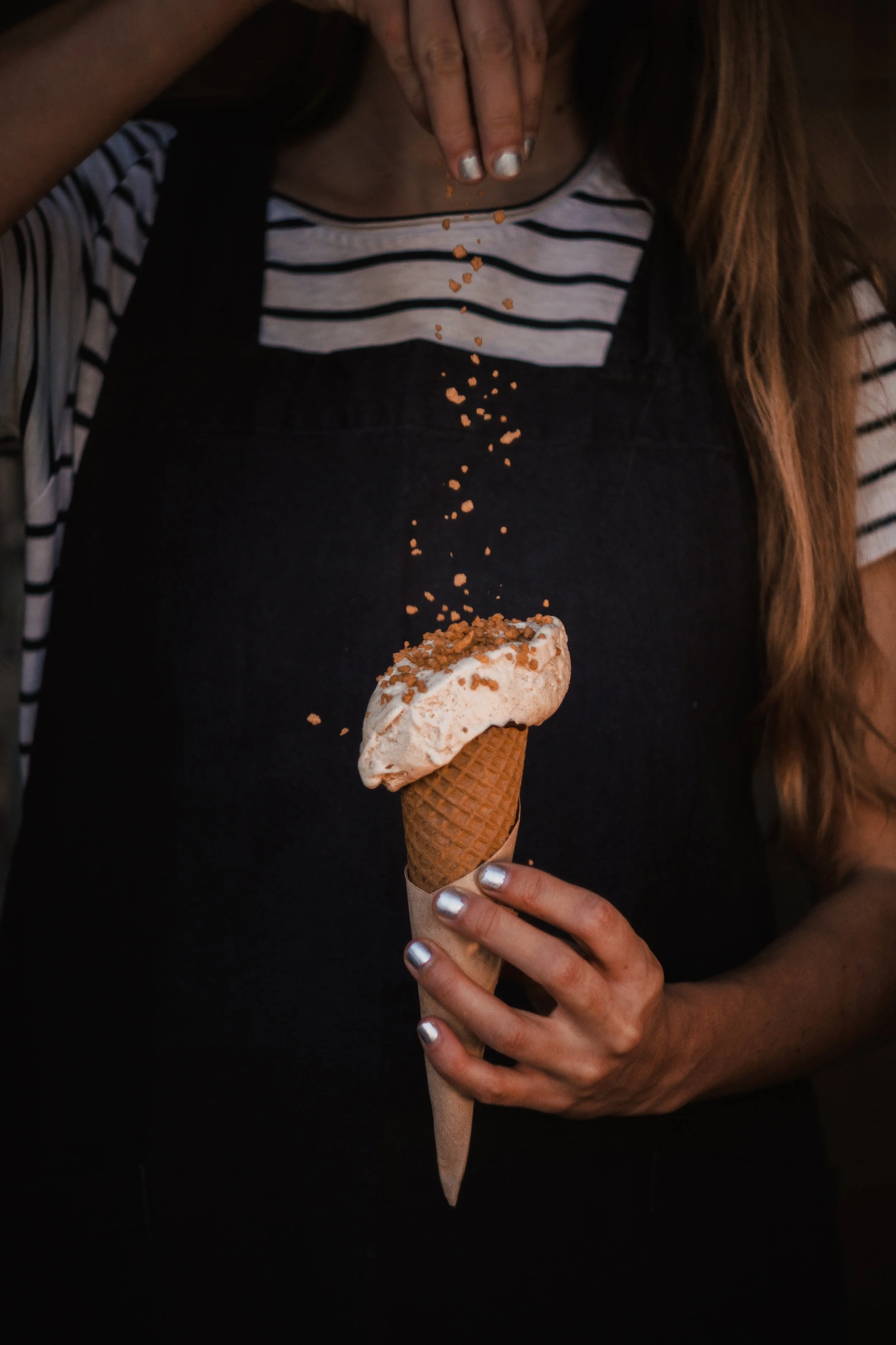 A person holding an ice cream cone with a scoop of ice cream topped with sprinkles, wearing a black and white striped shirt, with long brown hair.