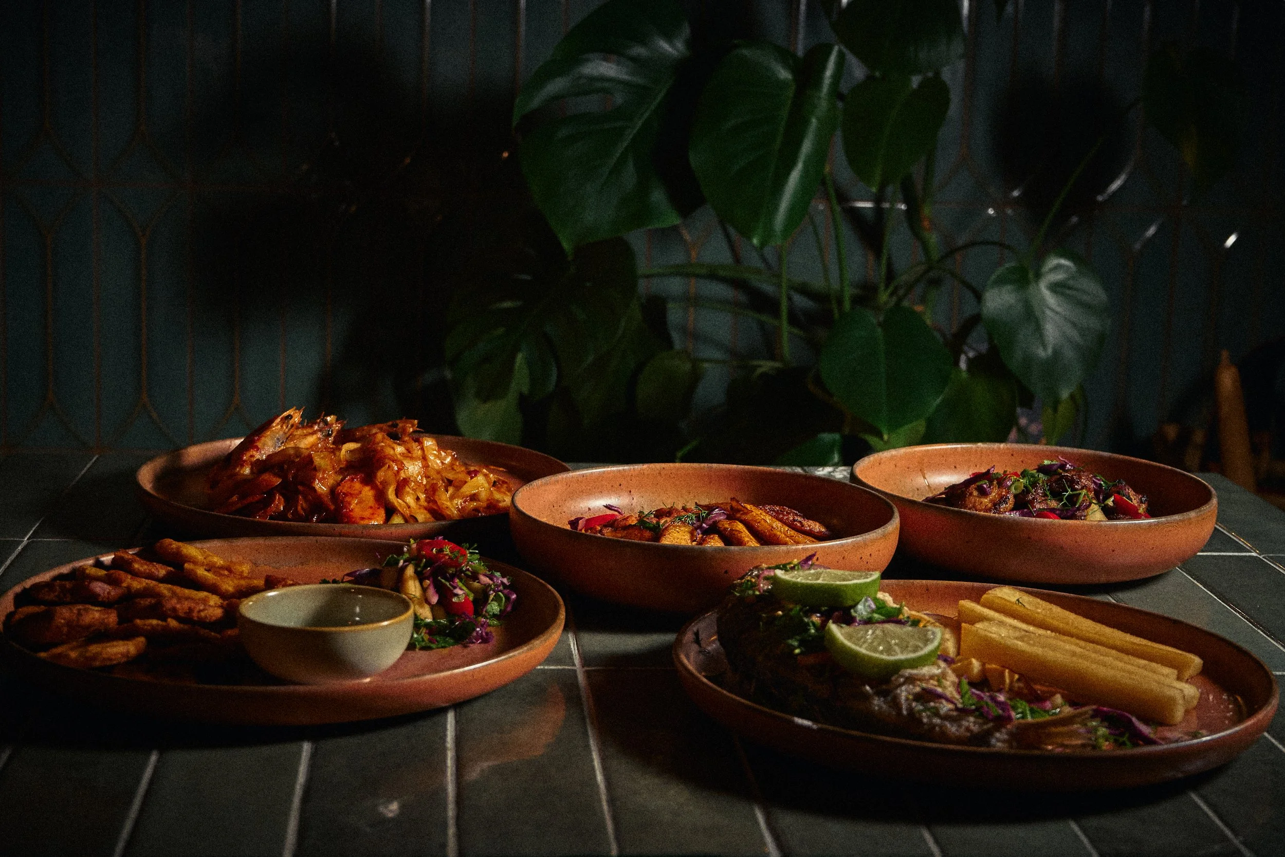 Various dishes of food arranged on a dark table with a large green leafy plant in the background.