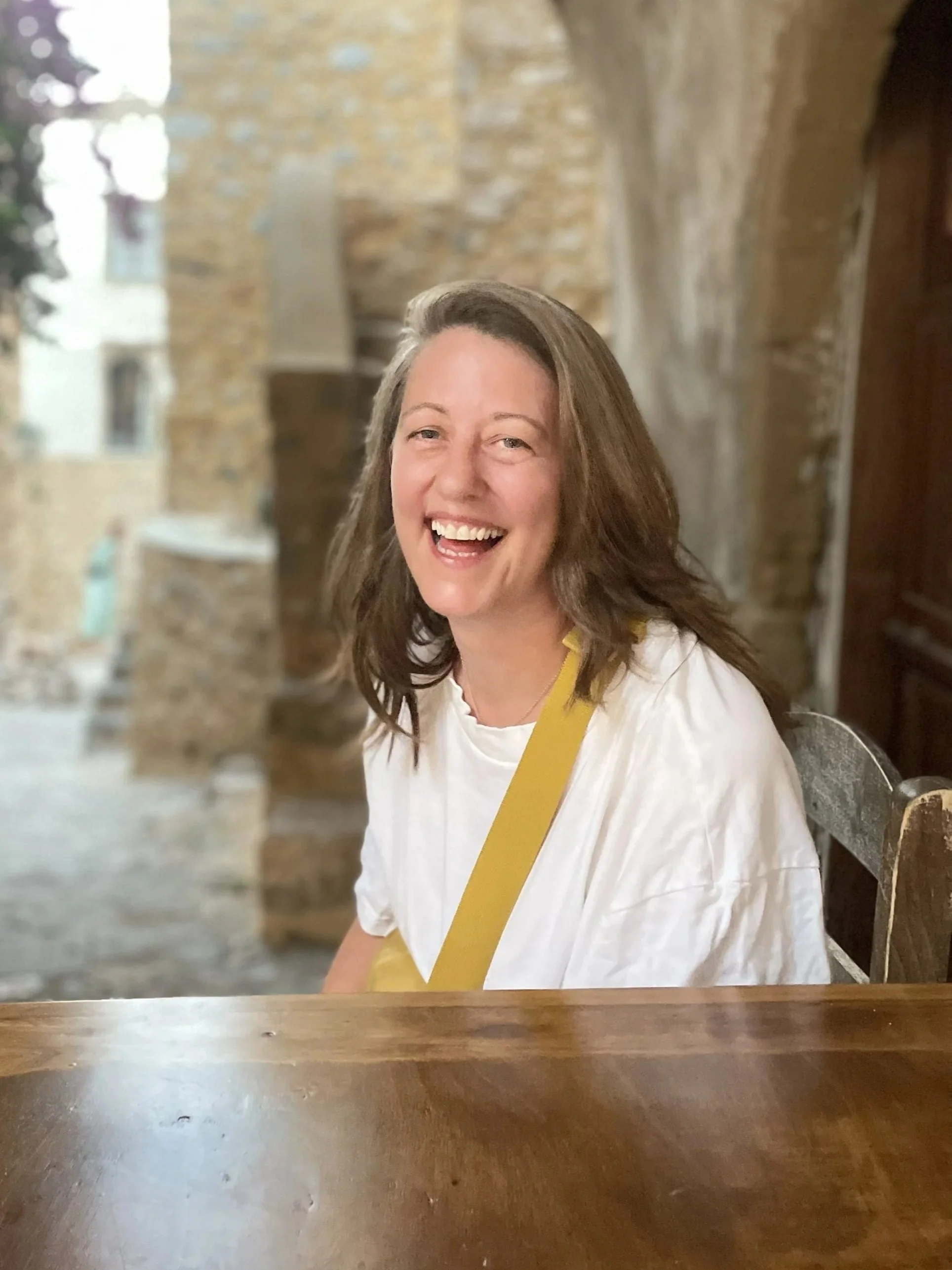 A woman with long brown hair smiling while sitting at a wooden table, wearing a white shirt and yellow strap, in a rustic setting with exposed brick walls and blurred background.