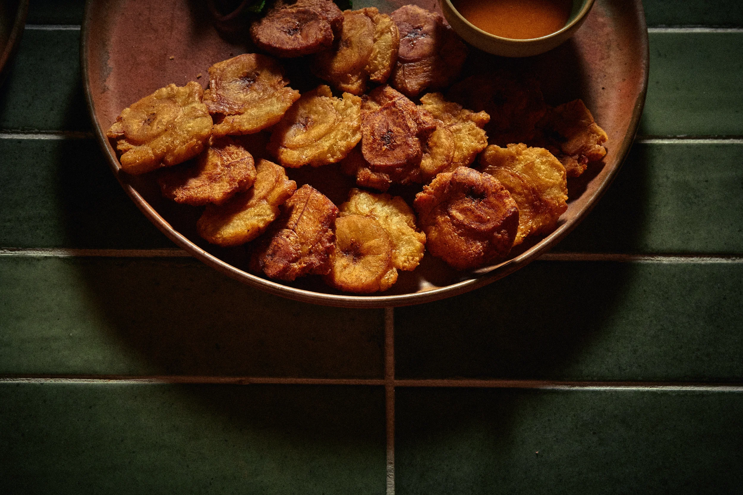A plate of fried plantains and fried chicken pieces with a small bowl of dipping sauce on a green tiled surface.
