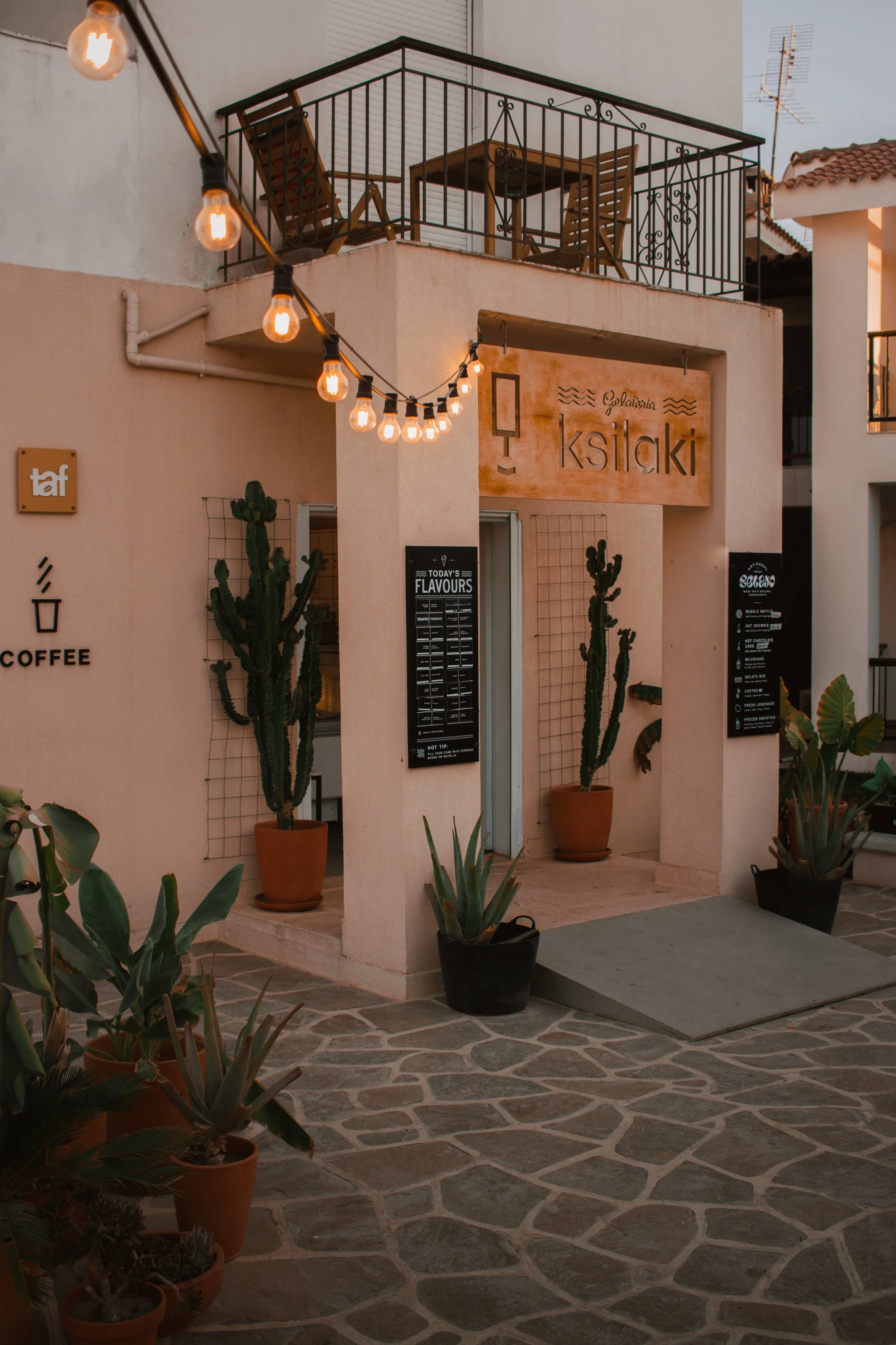 A cozy outdoor patio area with a stone floor, potted cacti and succulents, string lights hanging, and a building with a sign that reads 'ksiaki'. The balcony has a small table and chairs, and a wall-mounted menu board is visible.