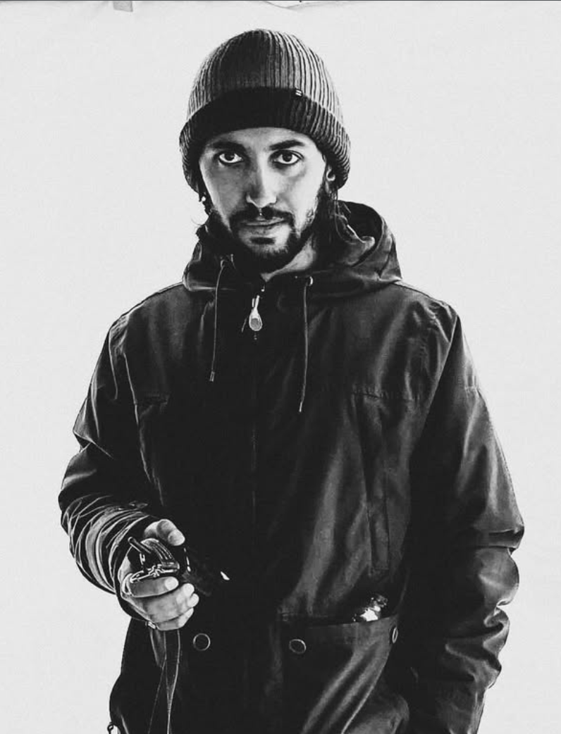 Black and white photo of a man wearing a knitted beanie, a zip-up jacket, and holding headphones and a phone, looking directly at the camera.