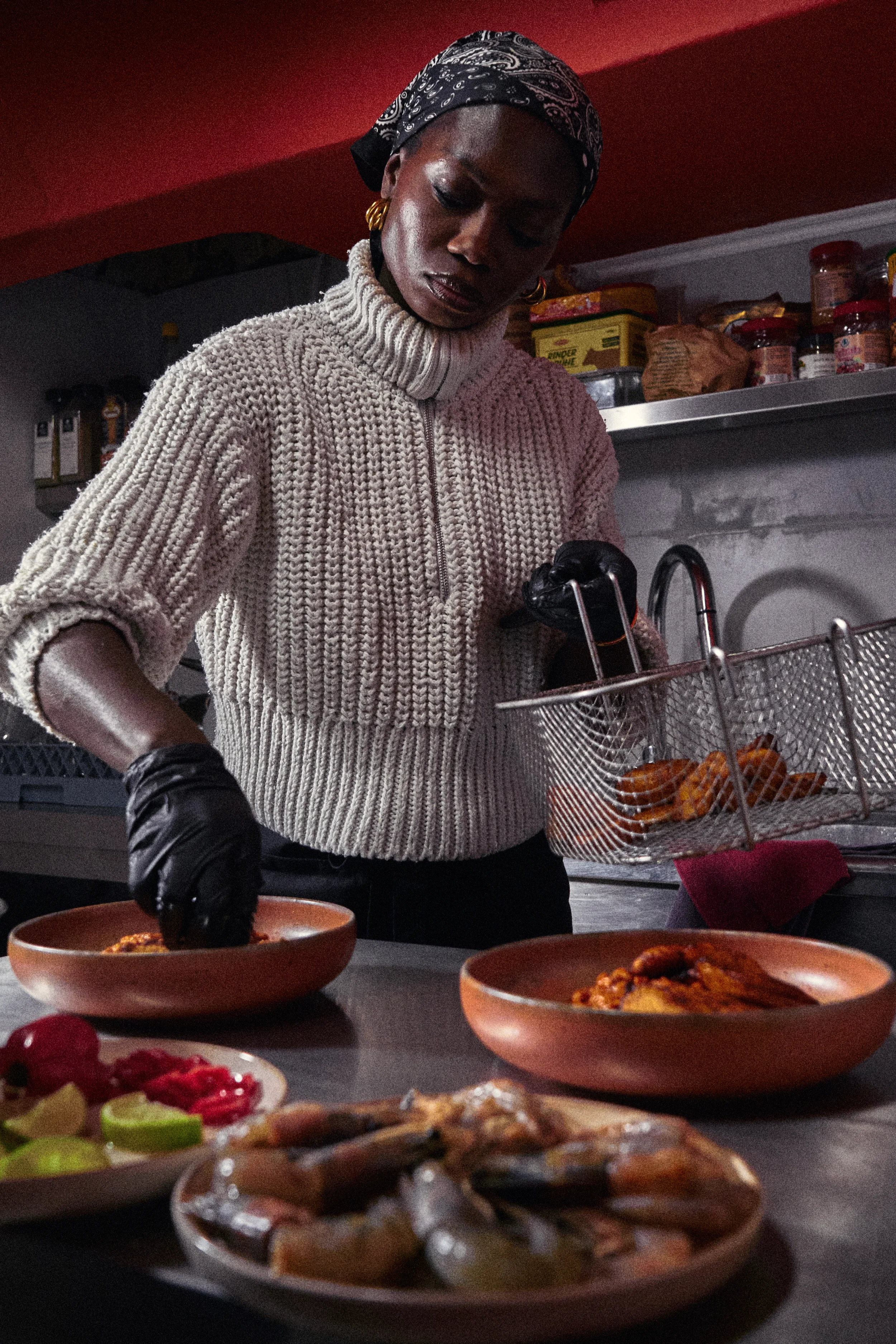 A woman wearing a beige knitted sweater, gold earrings, and a black-patterned headscarf prepares food in a kitchen. She is handling a basket of fried items, possibly chicken wings, over a table with bowls of food, including a bowl of red sauce and sliced vegetables. The kitchen background includes shelves with jars and boxes.