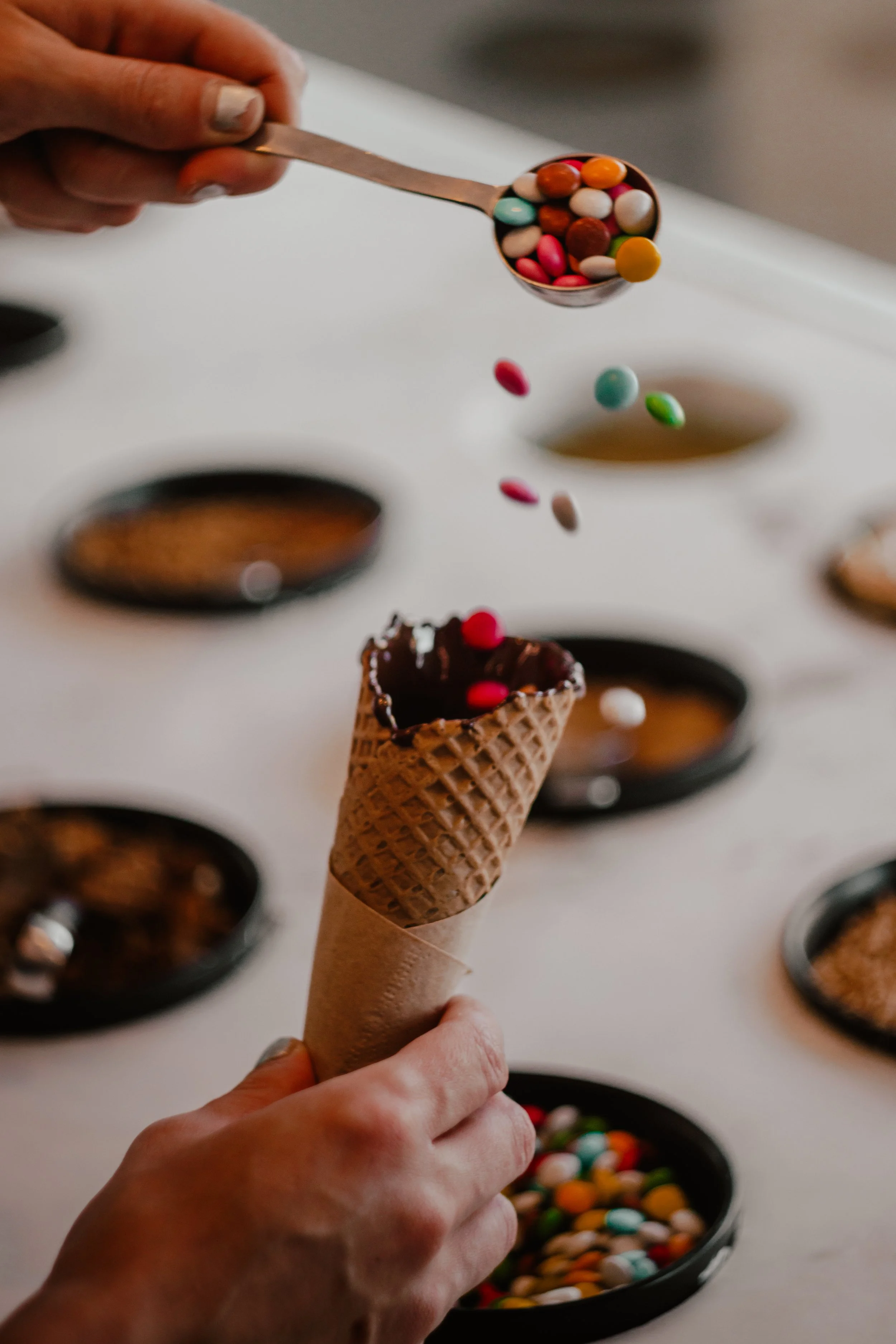 Person holding an ice cream cone with chocolate coating, filled with colorful candies, over bowls of candies on a white surface.