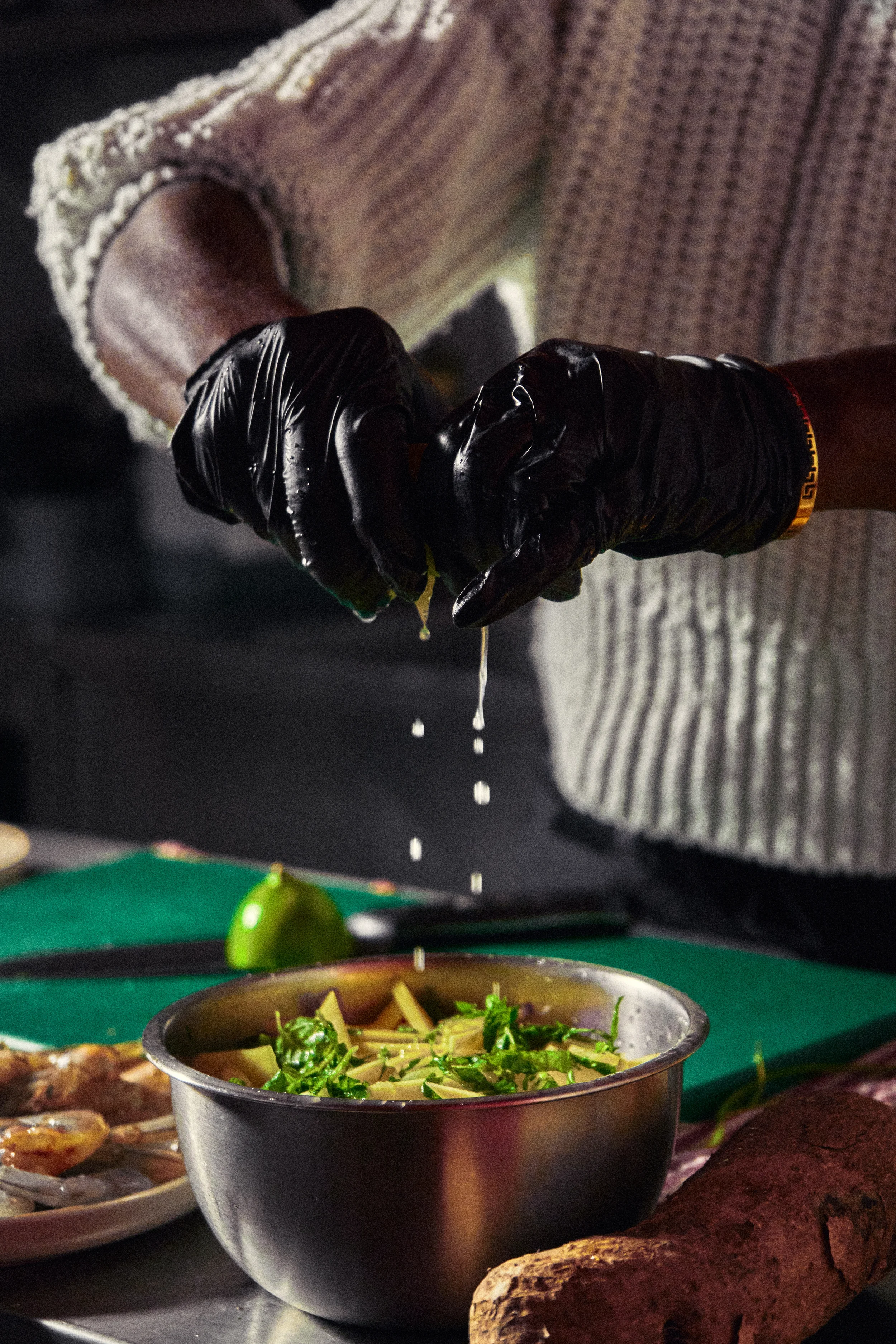 Person pouring a dish of noodles and vegetables from a small metal cup onto a table with other food items, including a baked sweet potato and a plate of grilled food, in a dimly lit setting.