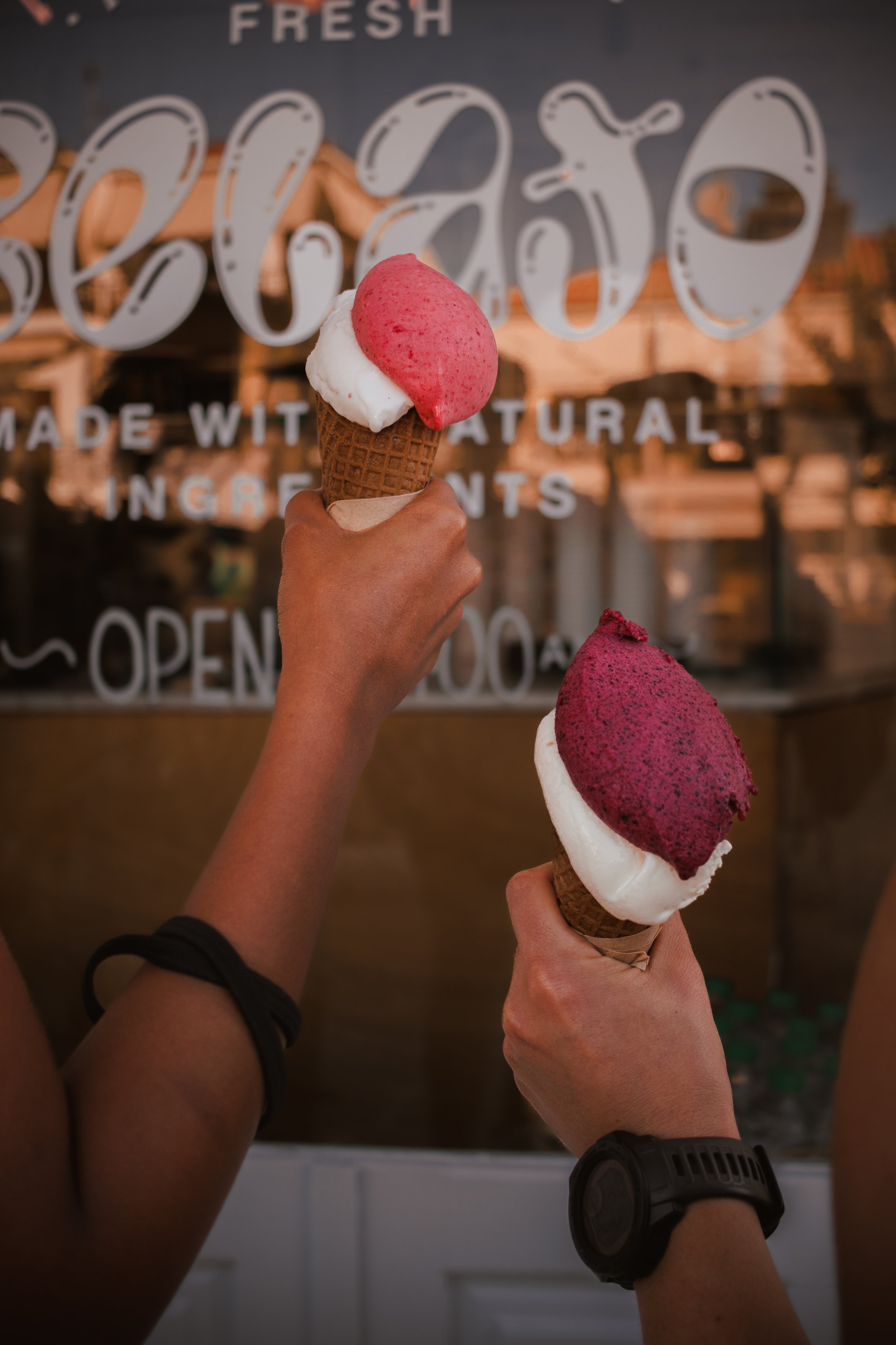 Two people holding ice cream cones, with a shop window in the background displaying the words 'FRESH' and 'made with natural ingredients'.