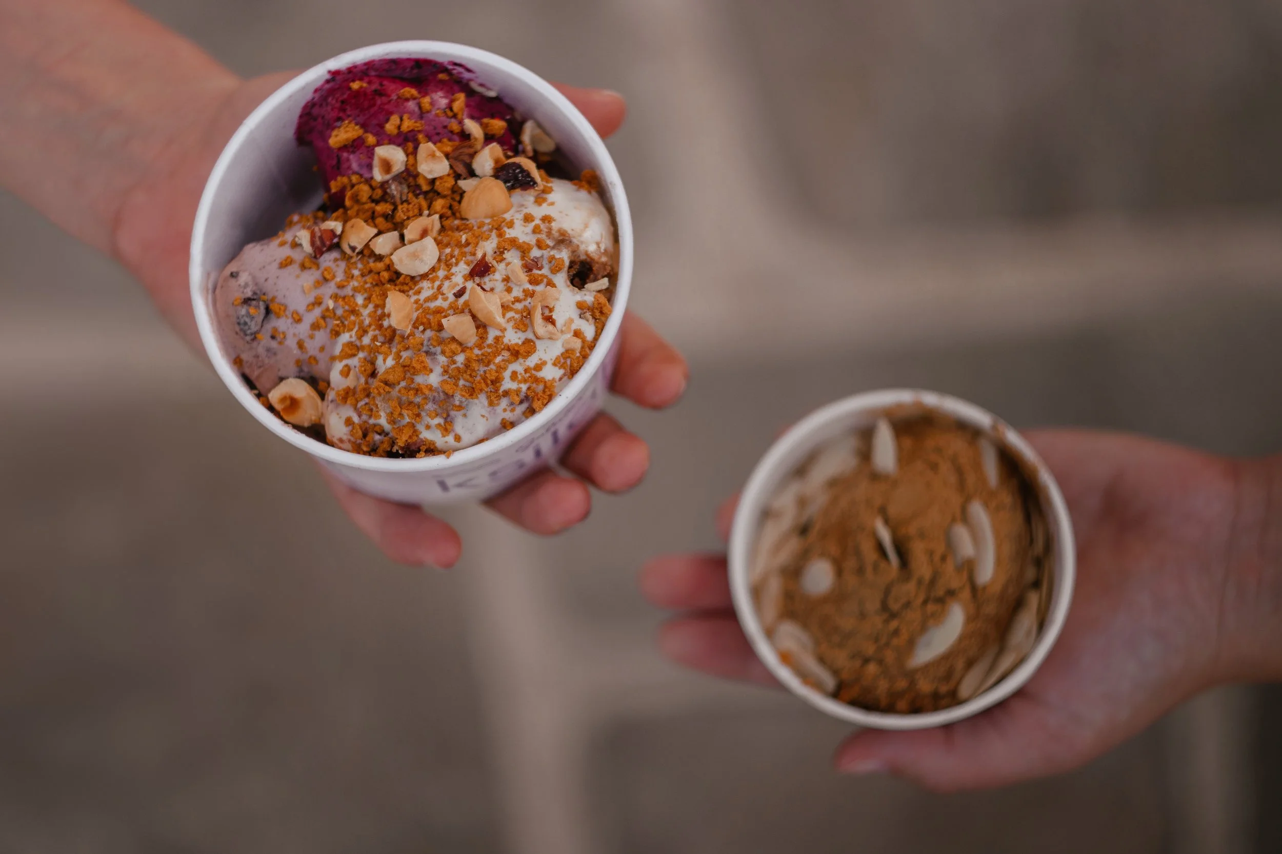 A person holding an ice cream cup topped with purple ice cream, chopped nuts, and crushed cookie crumbs. Another ice cream cup is visible in the background.