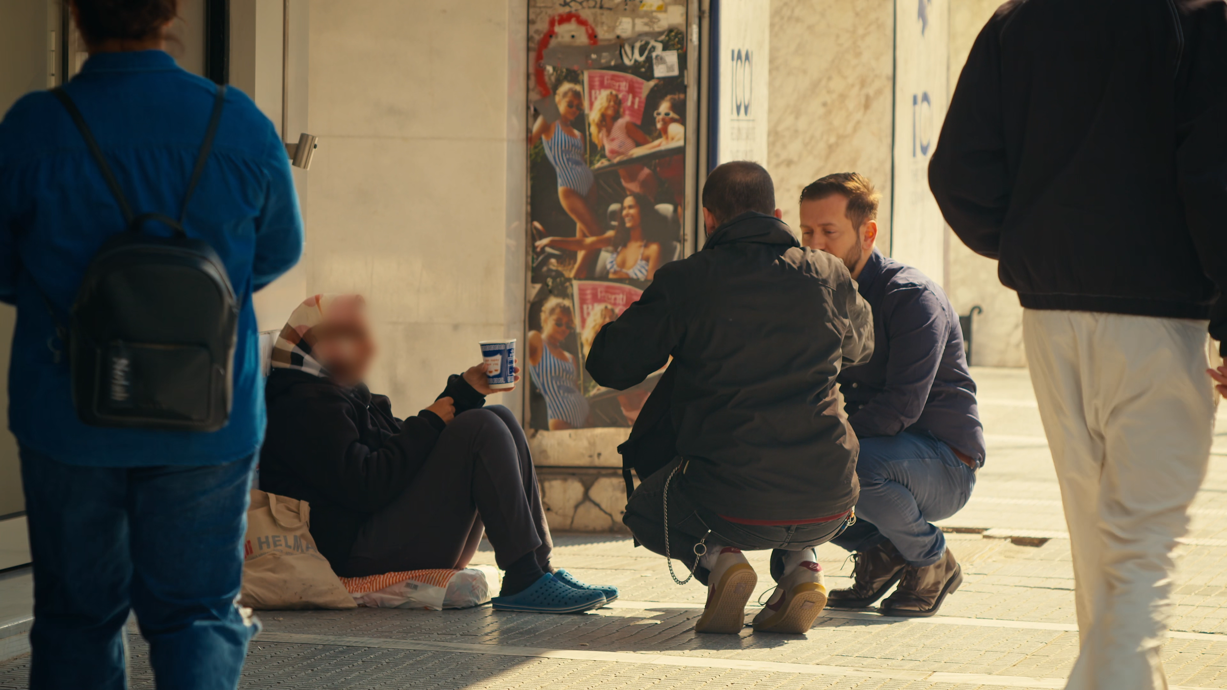 A group of people sitting and standing at a bus stop, some gathered and chatting, a woman holding a cup, and a few children nearby.