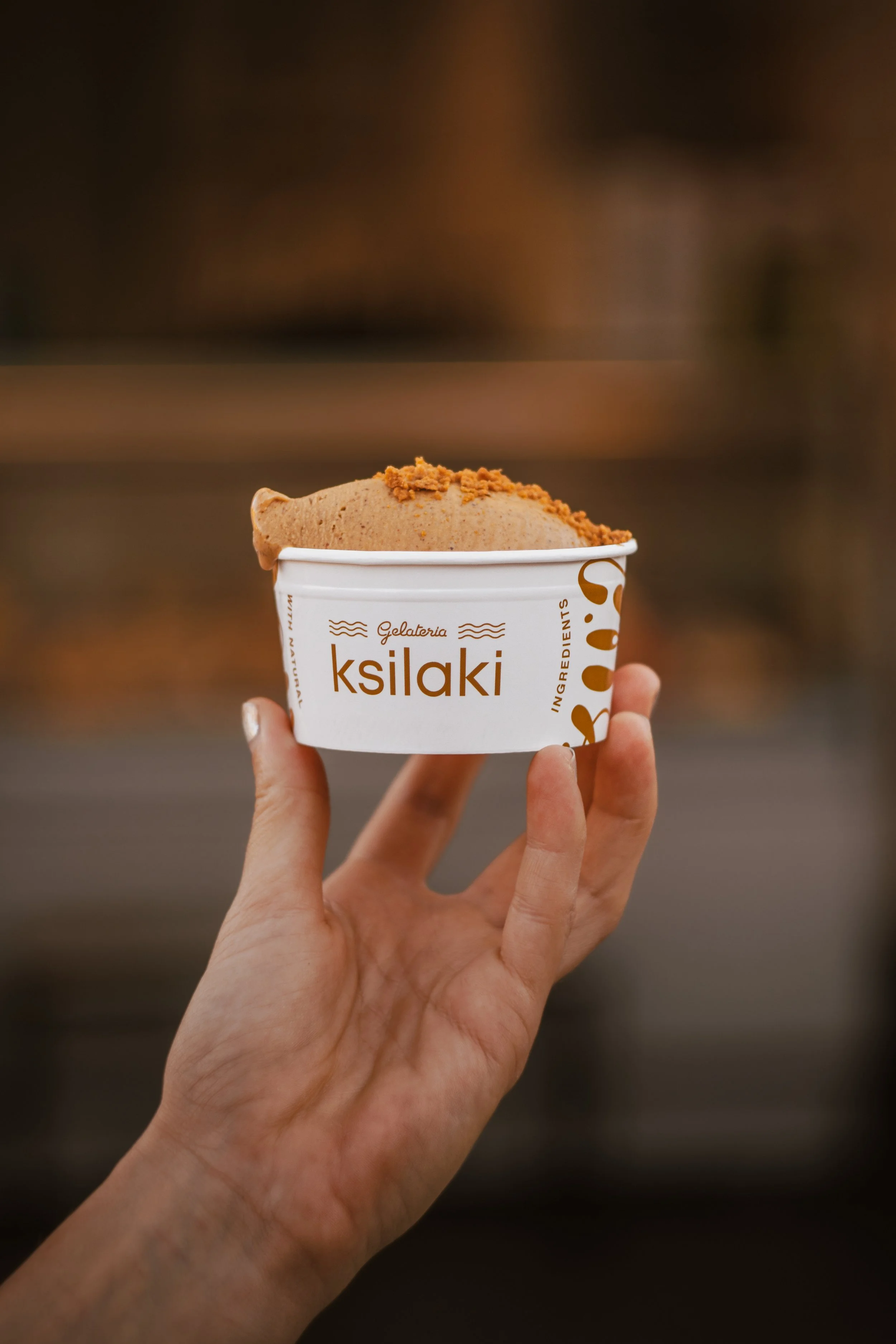 Hand holding a cup of gelato with a brown topping from a brand called ksilaki in front of a blurred background.