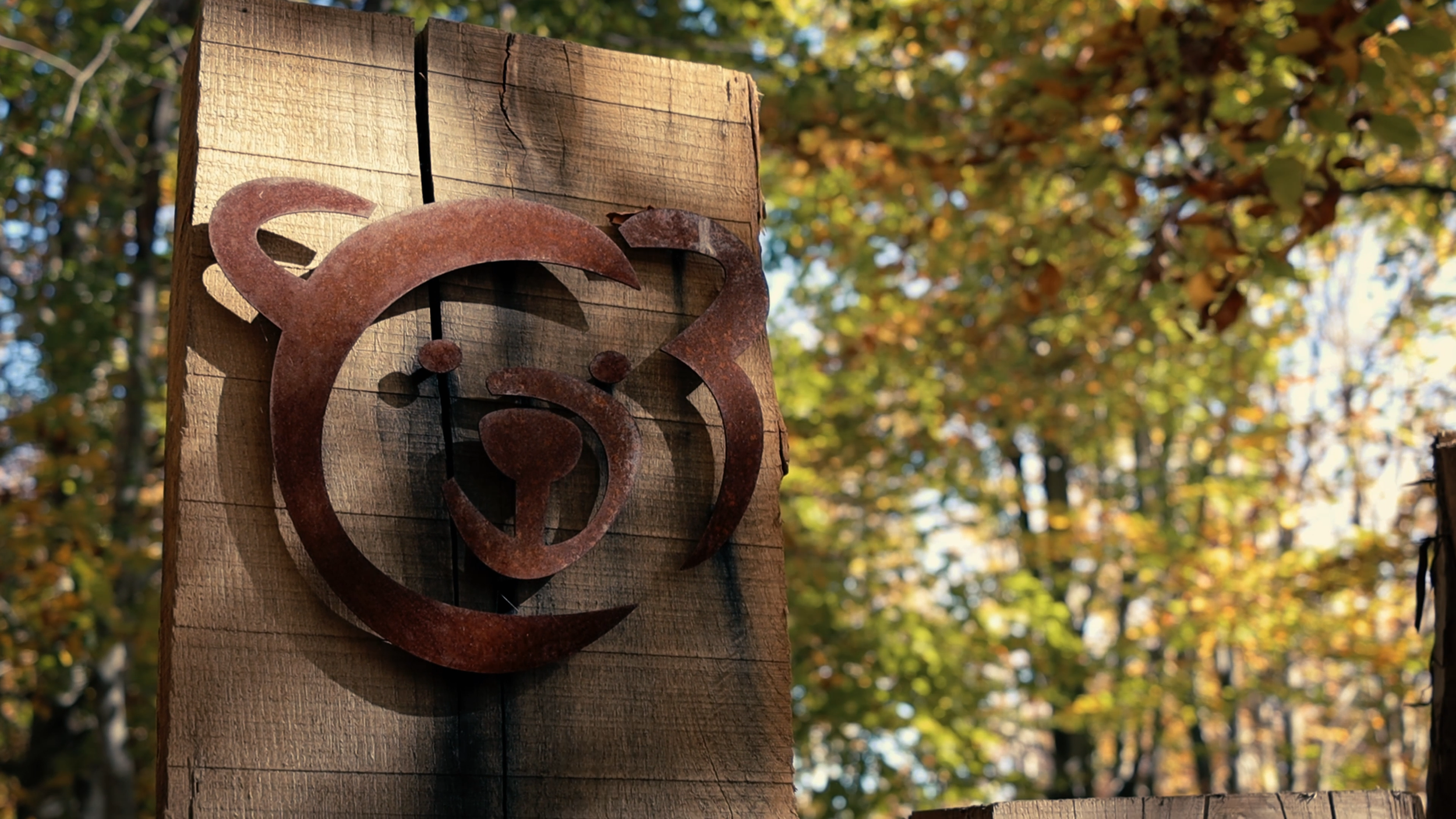Rusty metal heart and house-shaped decoration mounted on a weathered wooden post, with a background of trees with autumn leaves.