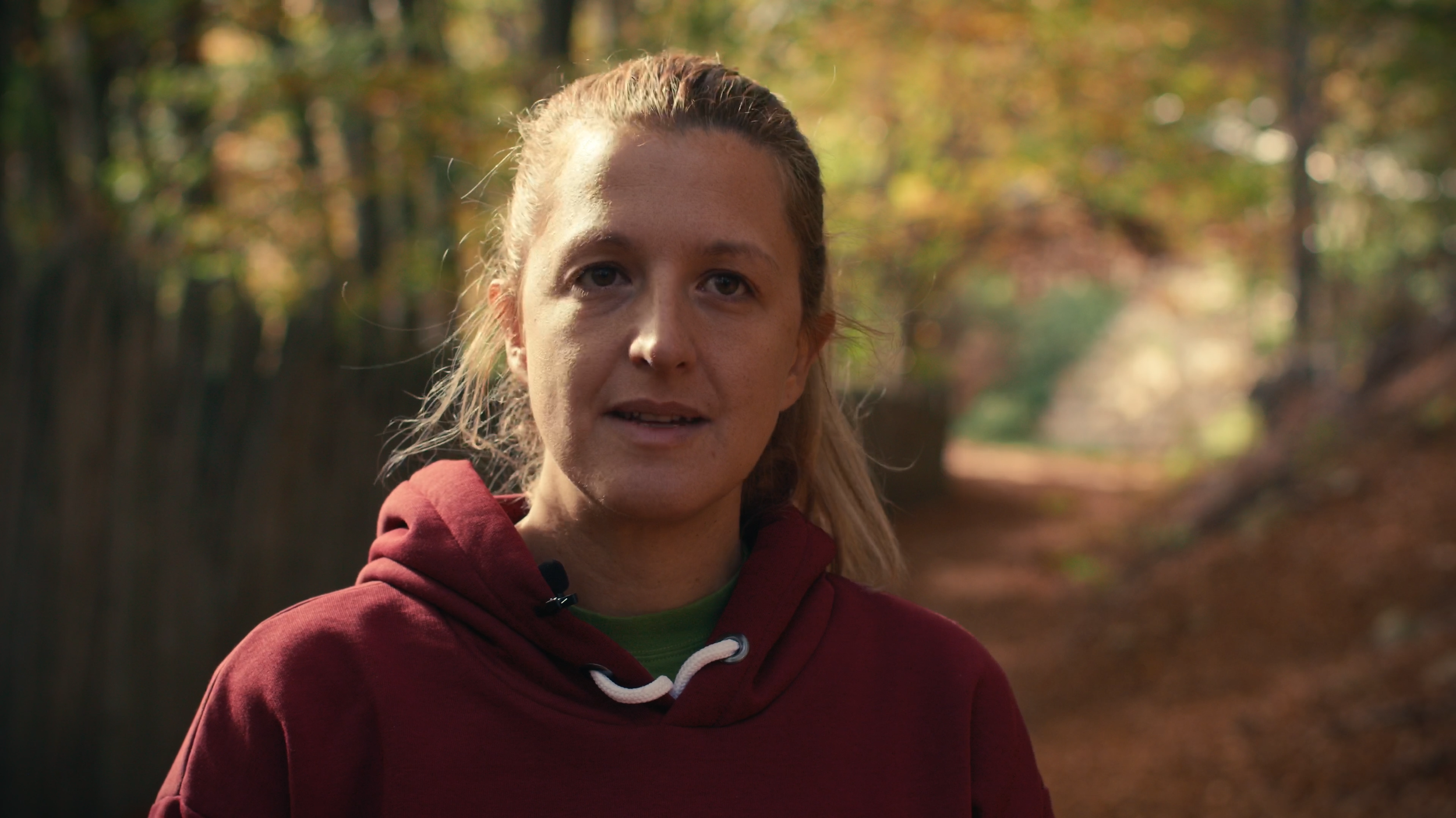 A woman with blonde hair wearing a red hoodie, standing outdoors in a wooded area with autumn foliage.