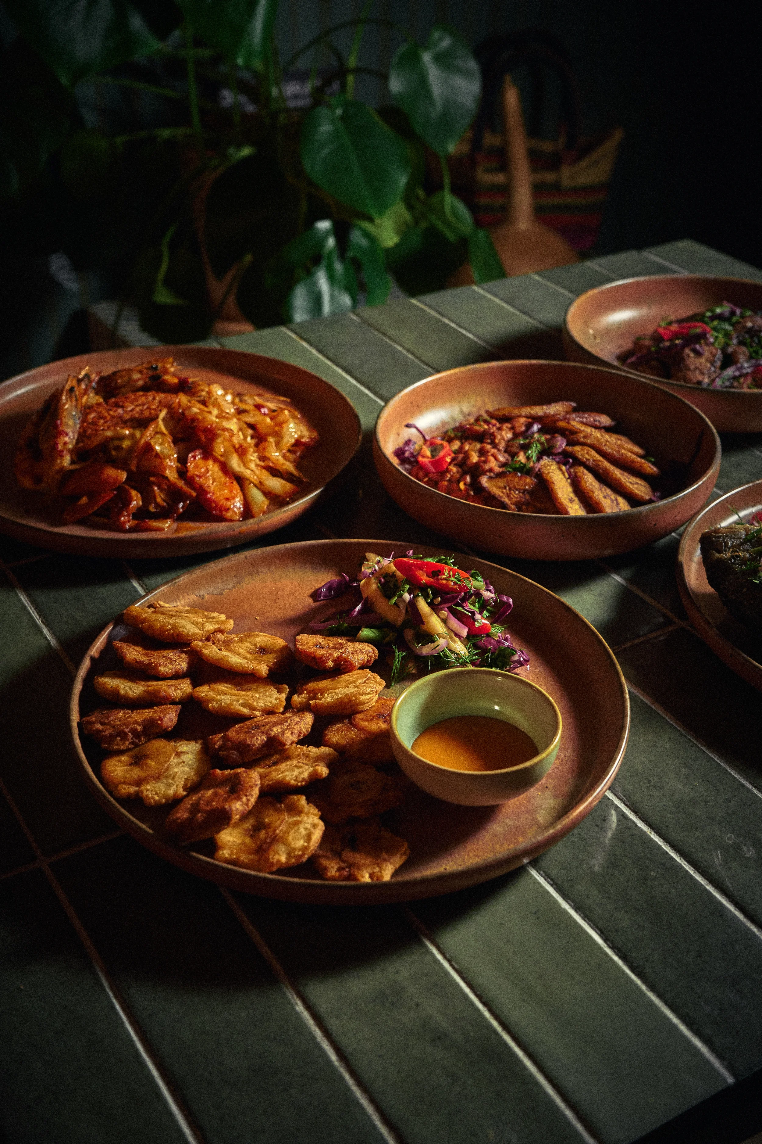 Several plates of food on a dark table, including fried plantain, grilled vegetables, and a salad, with a small cup of dipping sauce at the front.