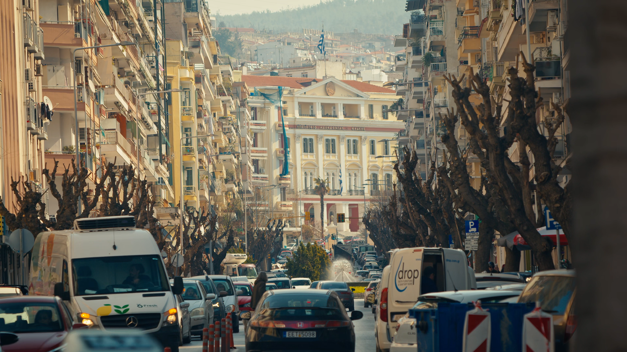 A busy city street filled with parked cars, moving vehicles, and tall buildings on either side. A historic building with Greek flags is visible in the background.