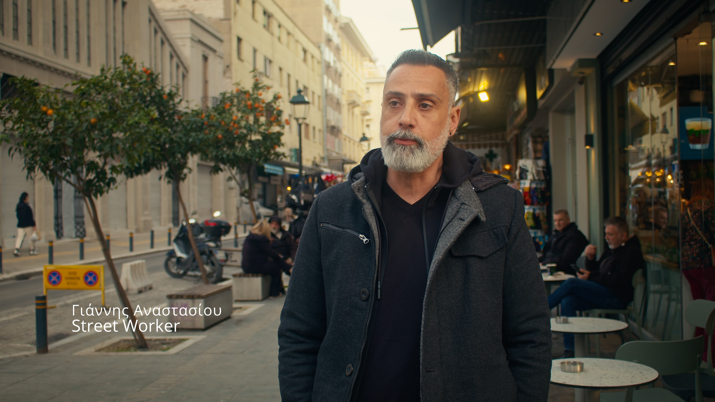 A middle-aged man with a gray beard and mustache, wearing a dark jacket and standing on a city street with shops, trees, and pedestrians in the background.