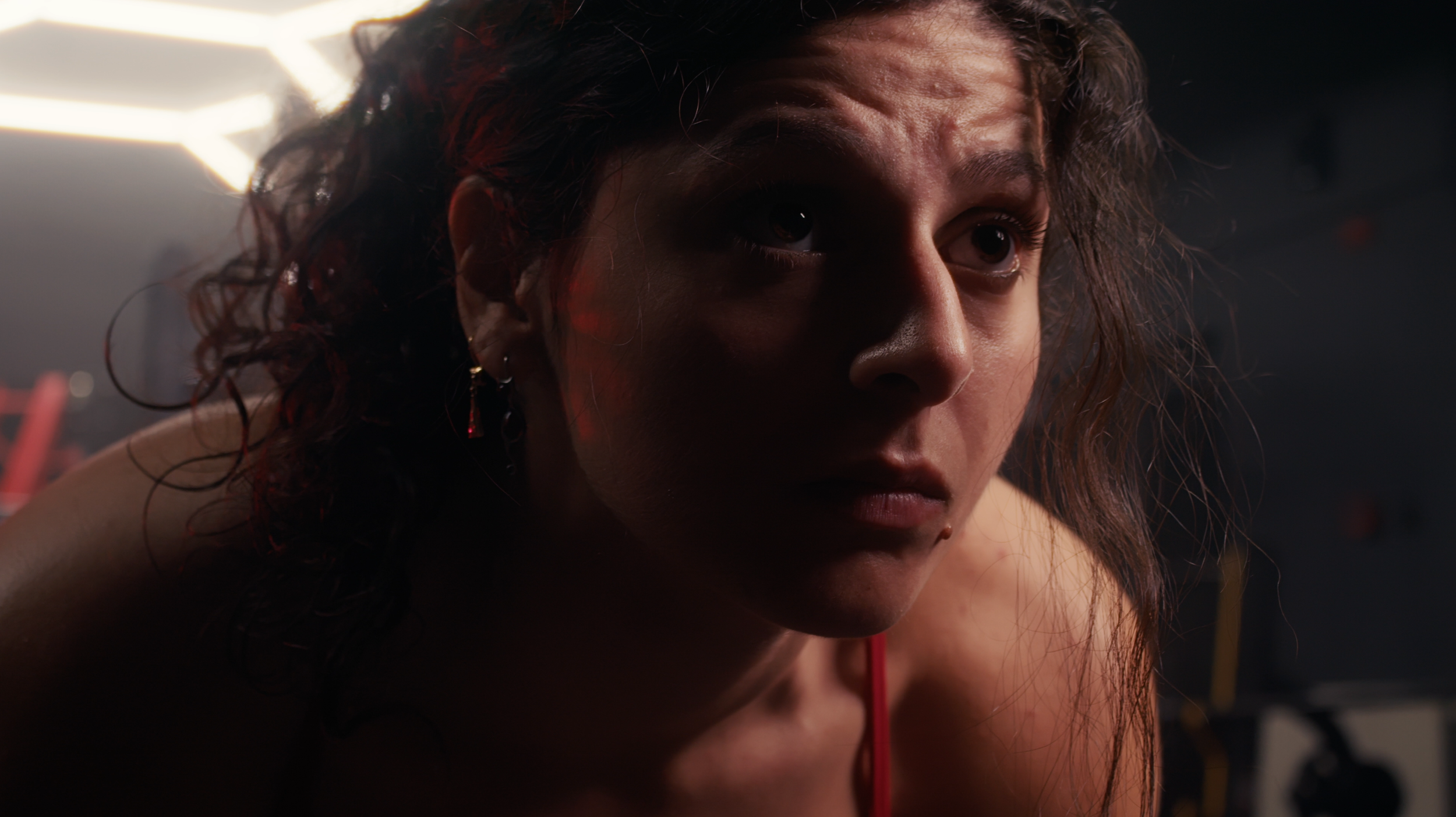 Close-up of a woman with curly hair and hoop earrings, looking intensely, with a dark background and warm lighting.