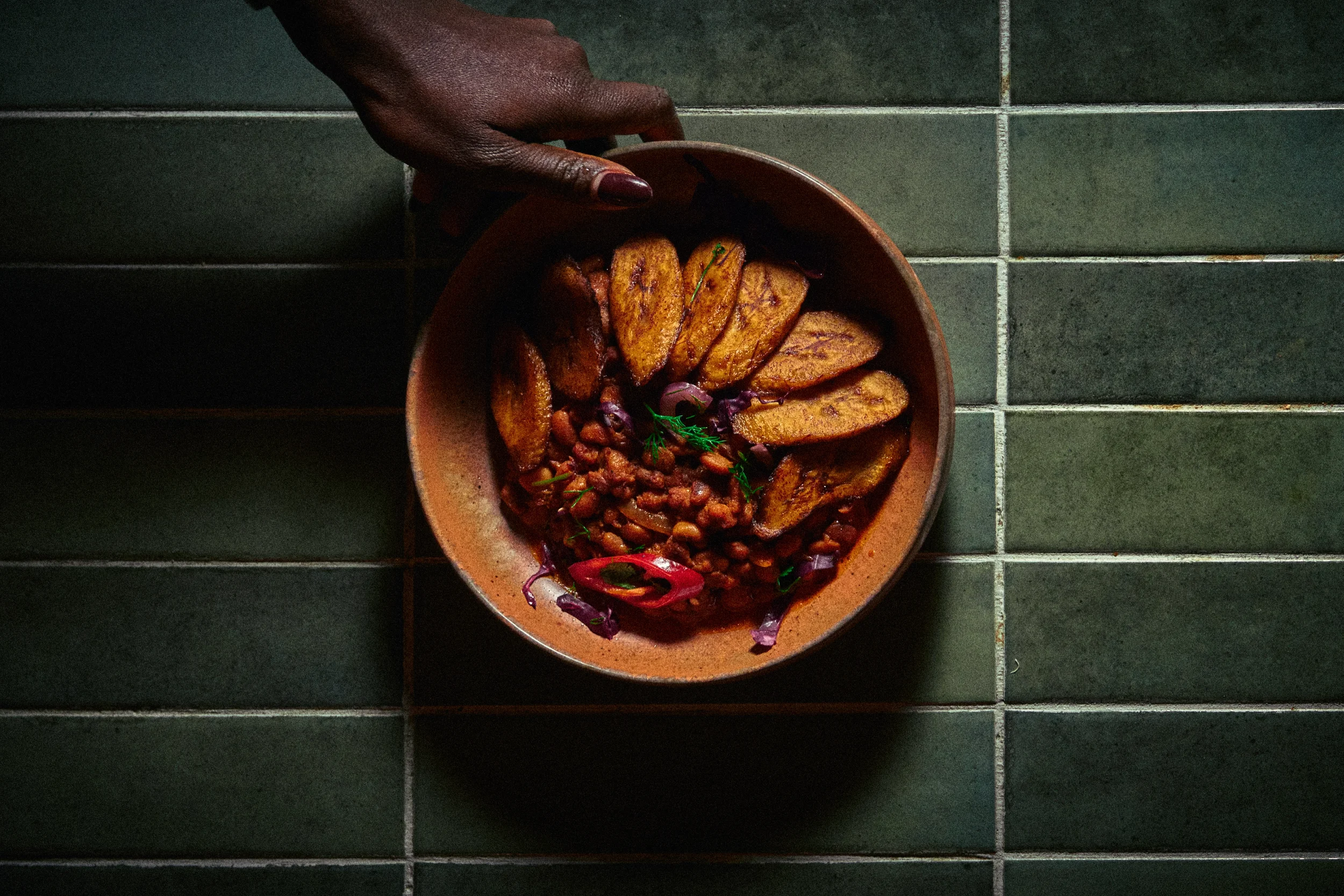A bowl of cooked plantains garnished with herbs, beans, and slices of red chili peppers, set on a dark green tiled surface.