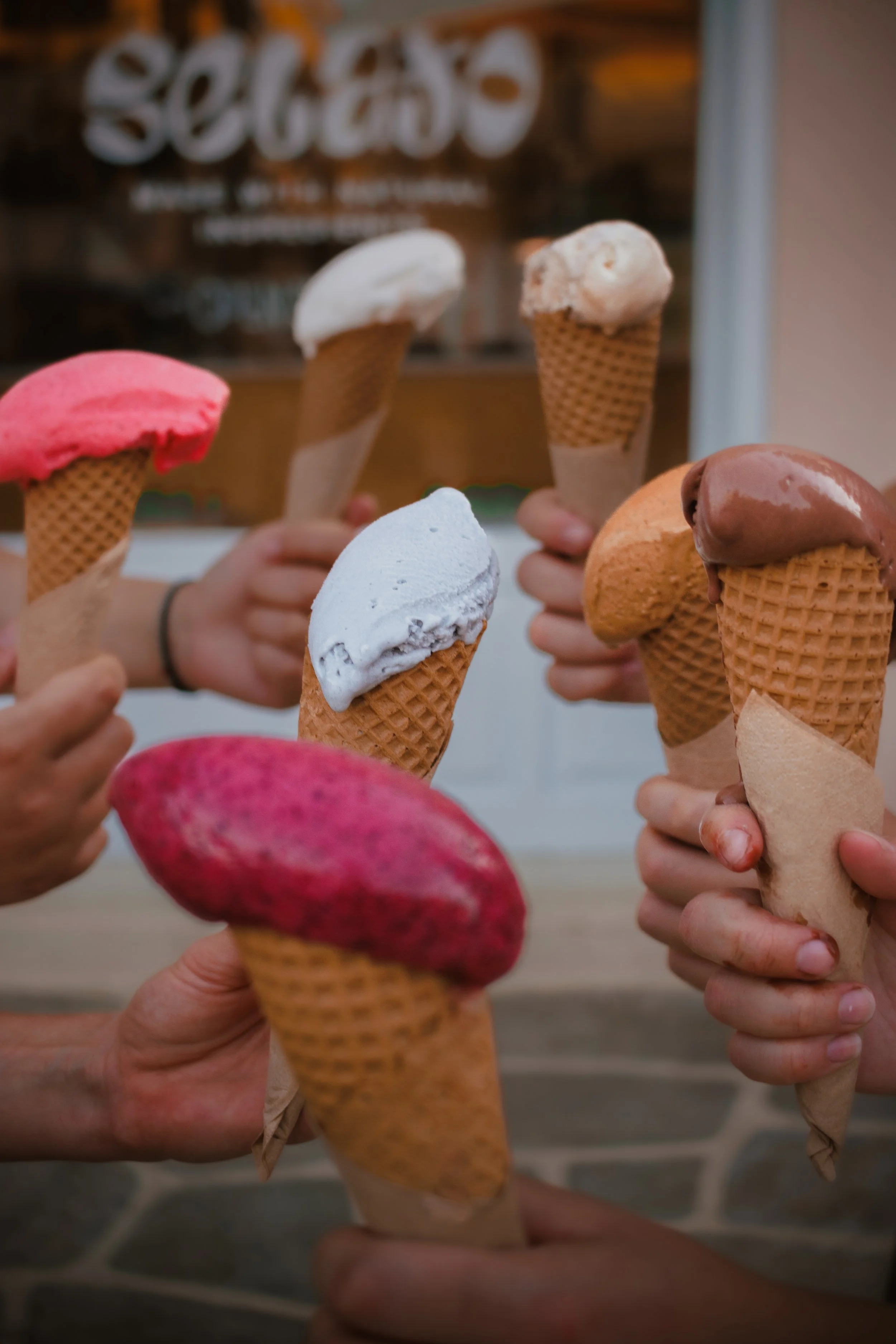 Multiple hands holding various colorful ice cream cones and an ice cream bar, with a blurred background featuring a sign that says 'gelato.'