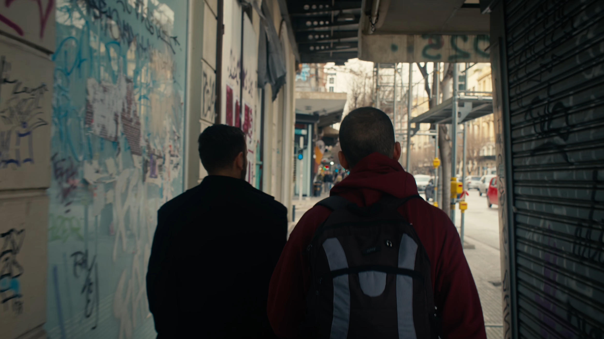 Two young men walking down a city sidewalk with graffiti on the wall to their left and storefronts on their right, with one wearing a red jacket and backpack and the other in darker clothing.