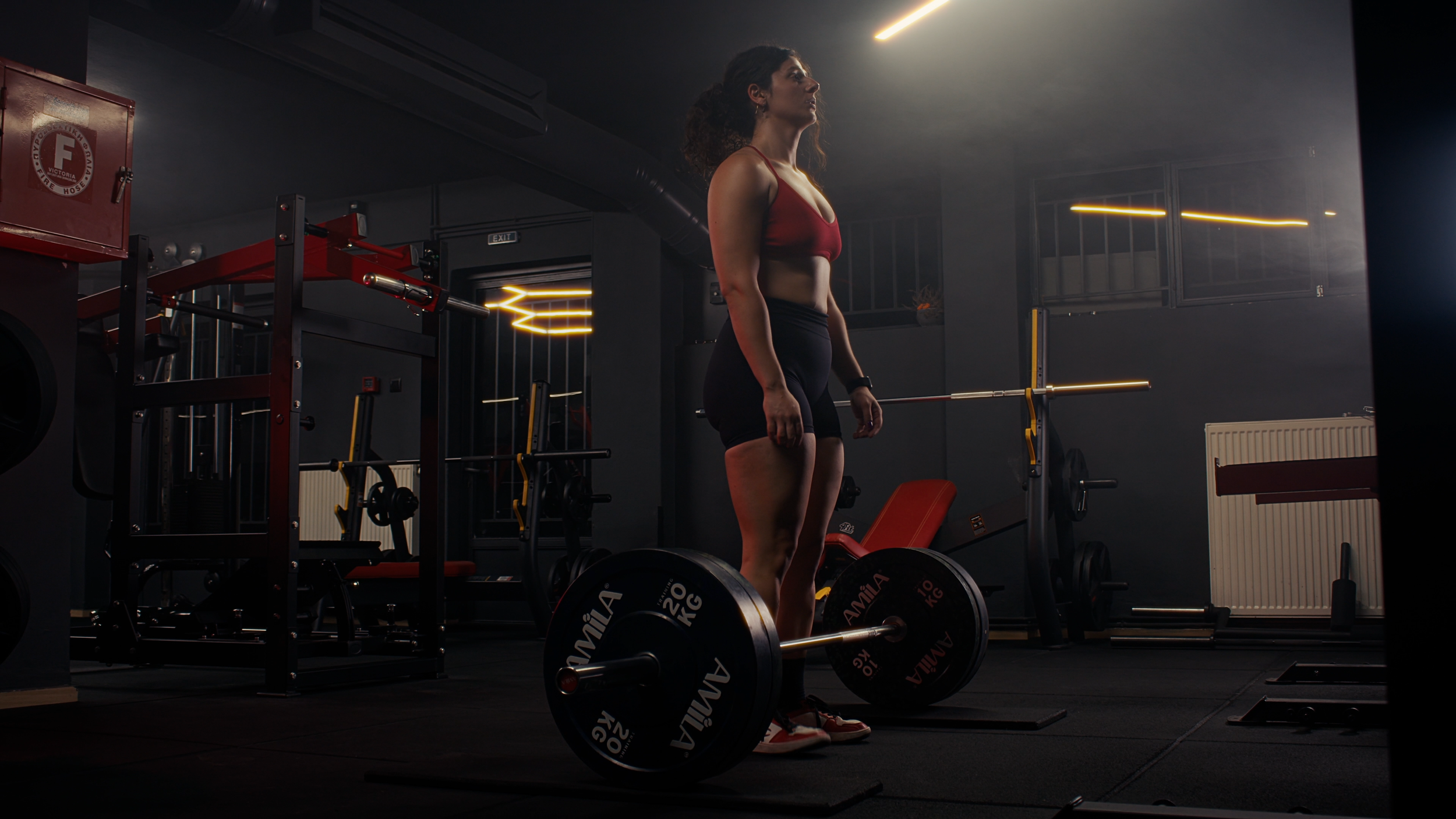 A woman in workout attire standing in a gym, preparing to lift a barbell with weight plates on the floor.