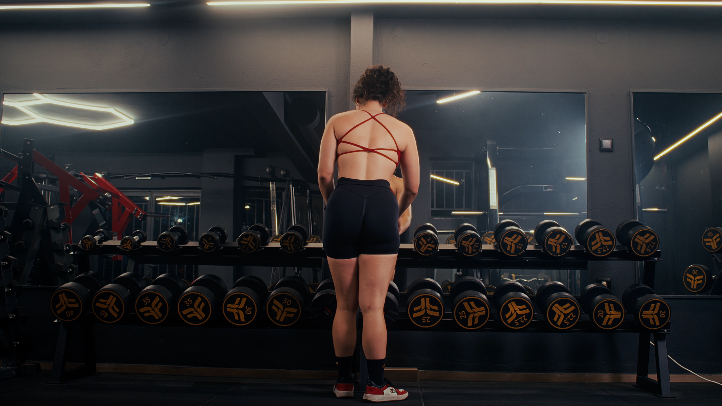 A woman standing in front of a mirror in a gym, wearing a crisscross back sports bra and shorts, looking at dumbbells arranged on a rack.
