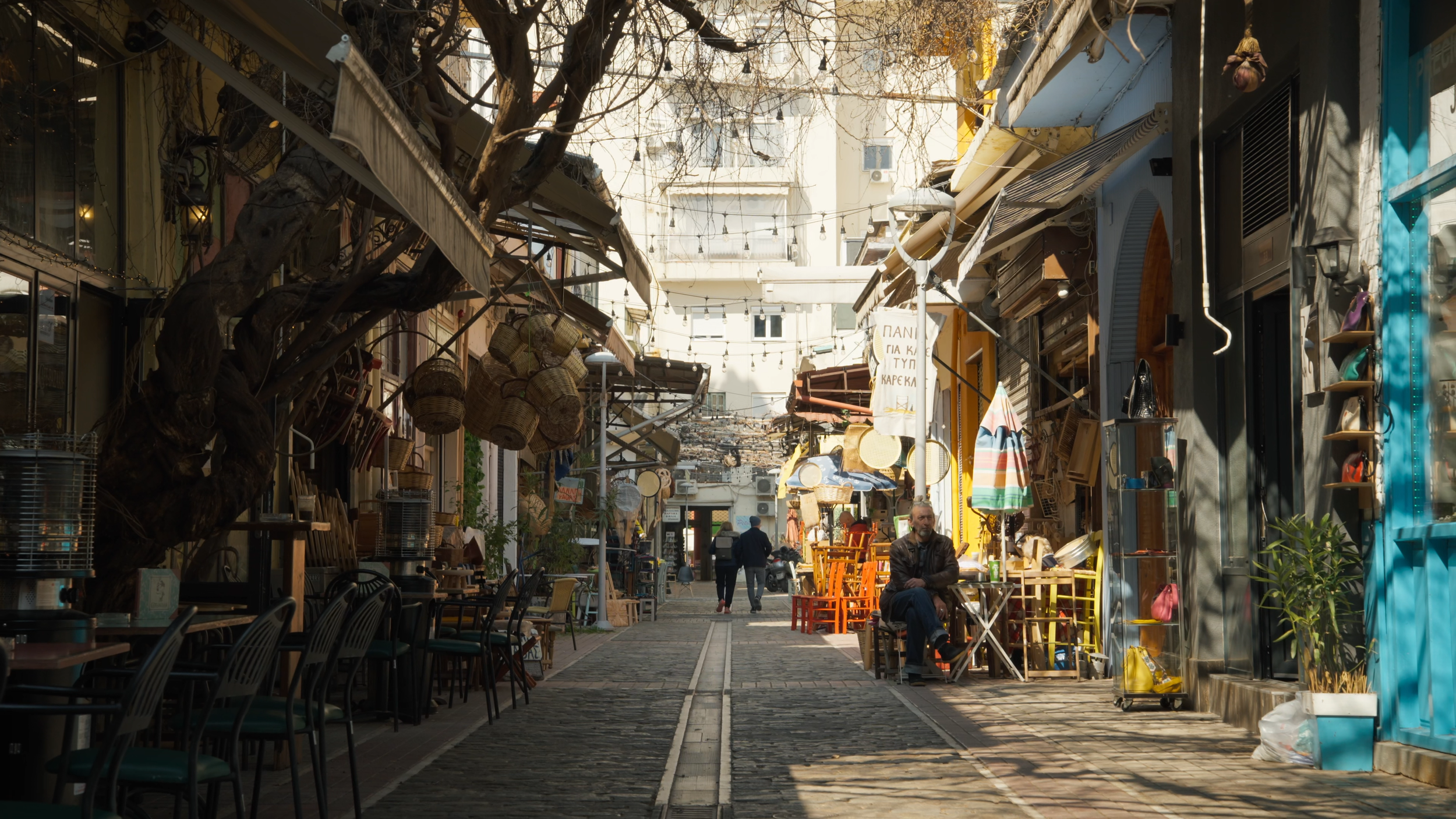 An outdoor marketplace with various shops and stands on a cobblestone street, decorated with hanging lights, with a large tree on the left and a man sitting on a chair in the foreground.