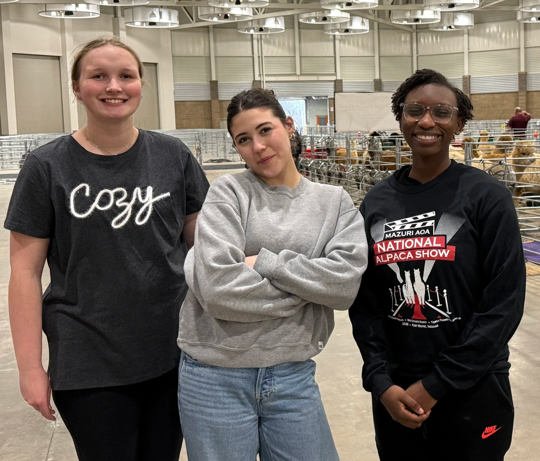 Three young women standing in an indoor space with metal pens containing alpacas in the background. All three are smiling, with the middle woman crossing her arms. The woman on the left has light skin and blonde hair, wearing a black t-shirt with white cursive writing. The woman in the middle has light skin, dark hair in a bun, and is wearing a gray sweatshirt. The woman on the right has dark skin, short dreadlocks, glasses, and a black t-shirt with a red and white design for the National Alpaca Show.