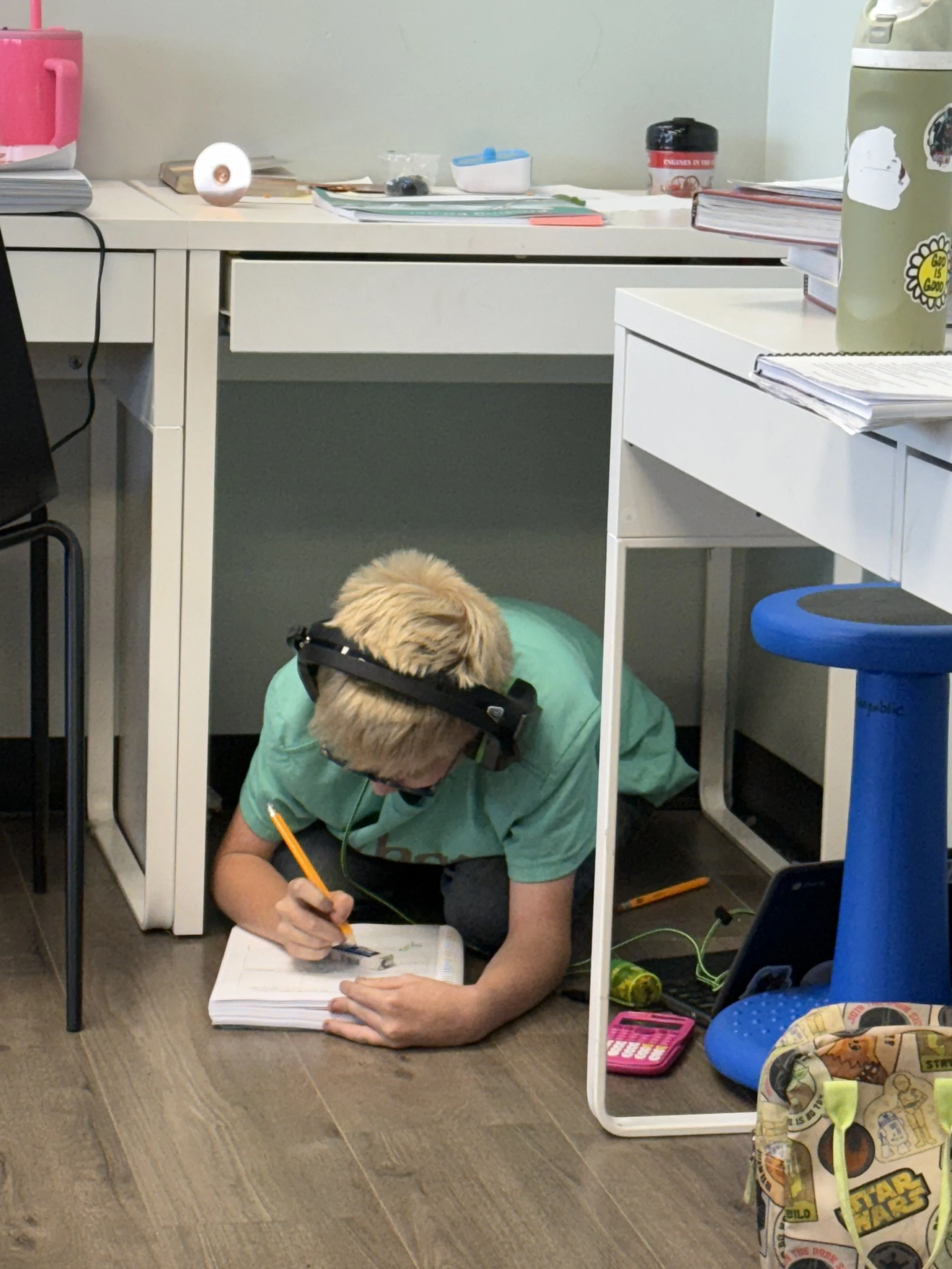 A person with blonde hair, wearing glasses and a green t-shirt, is working under a desk with a laptop and a notebook. The person is sitting on the floor and appears to be writing or drawing, with various office supplies scattered around, including a calculator, a toy, and a bag.