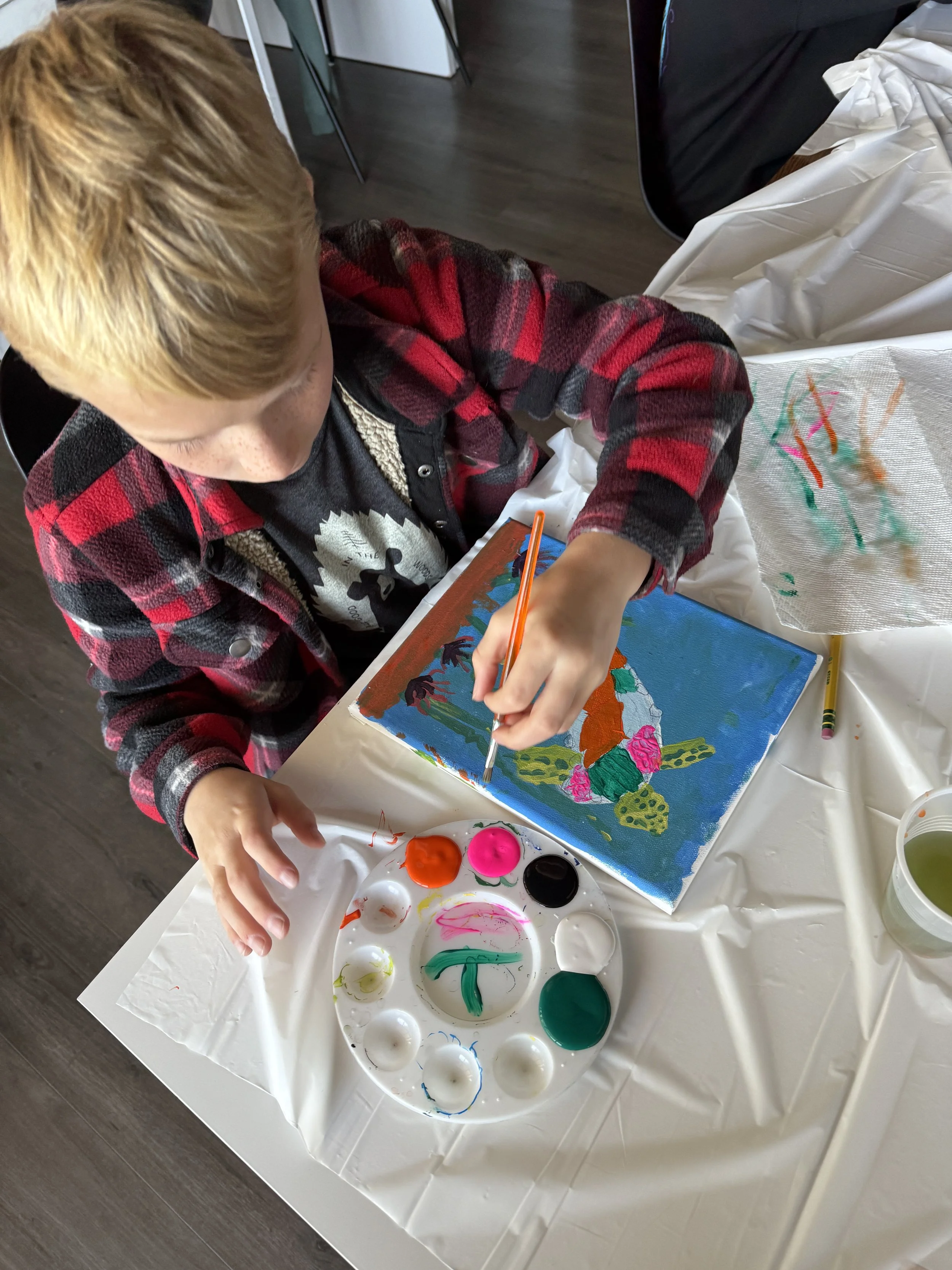 A person painting on a canvas with vibrant colors using a small brush, sitting at a table covered with a white plastic tablecloth and various pots of paint.