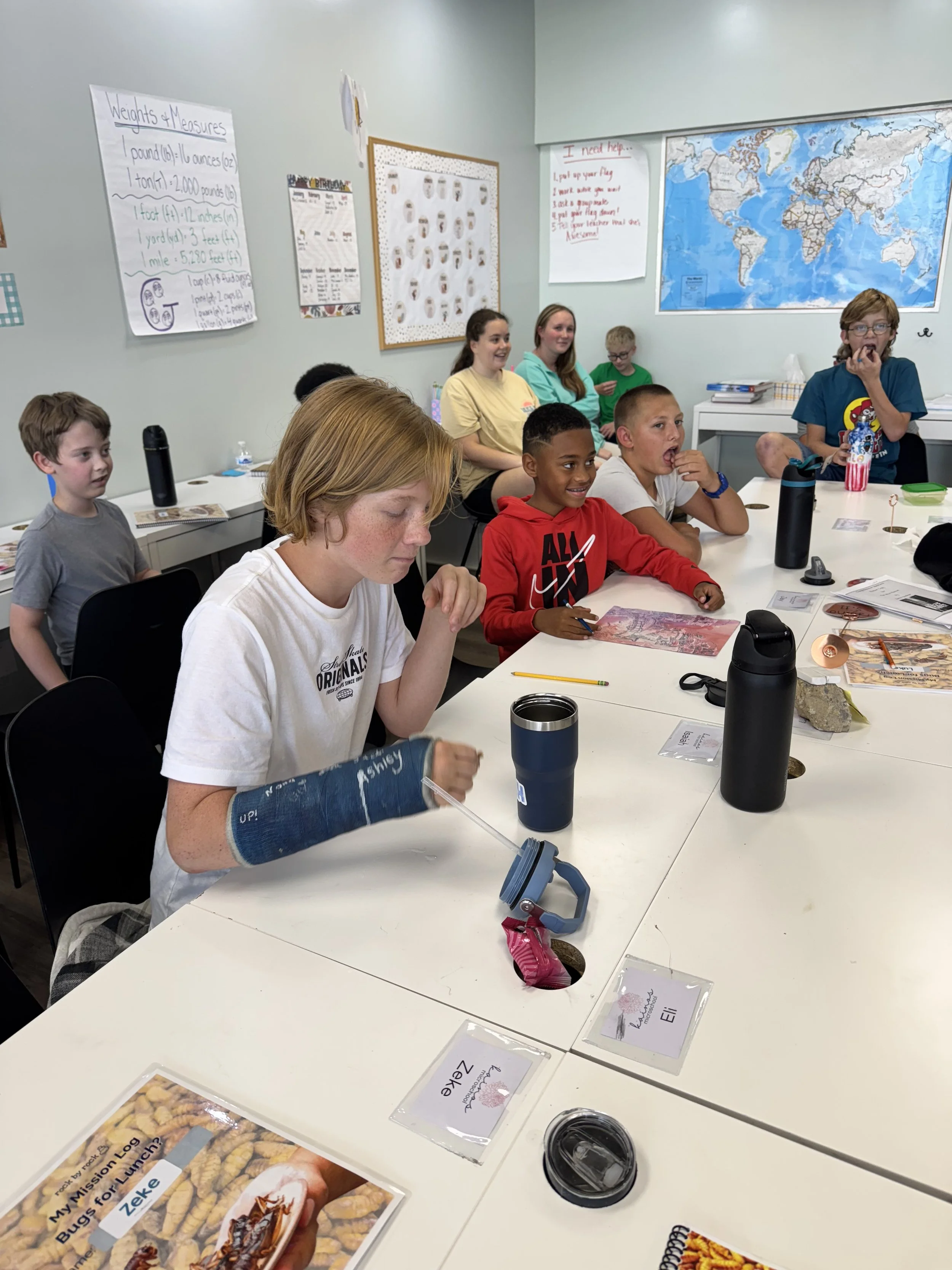 A classroom with children and teenagers sitting around a table, some smiling and engaging in activities. The table has name tags, books, and cups. Posters and a world map are on the walls.