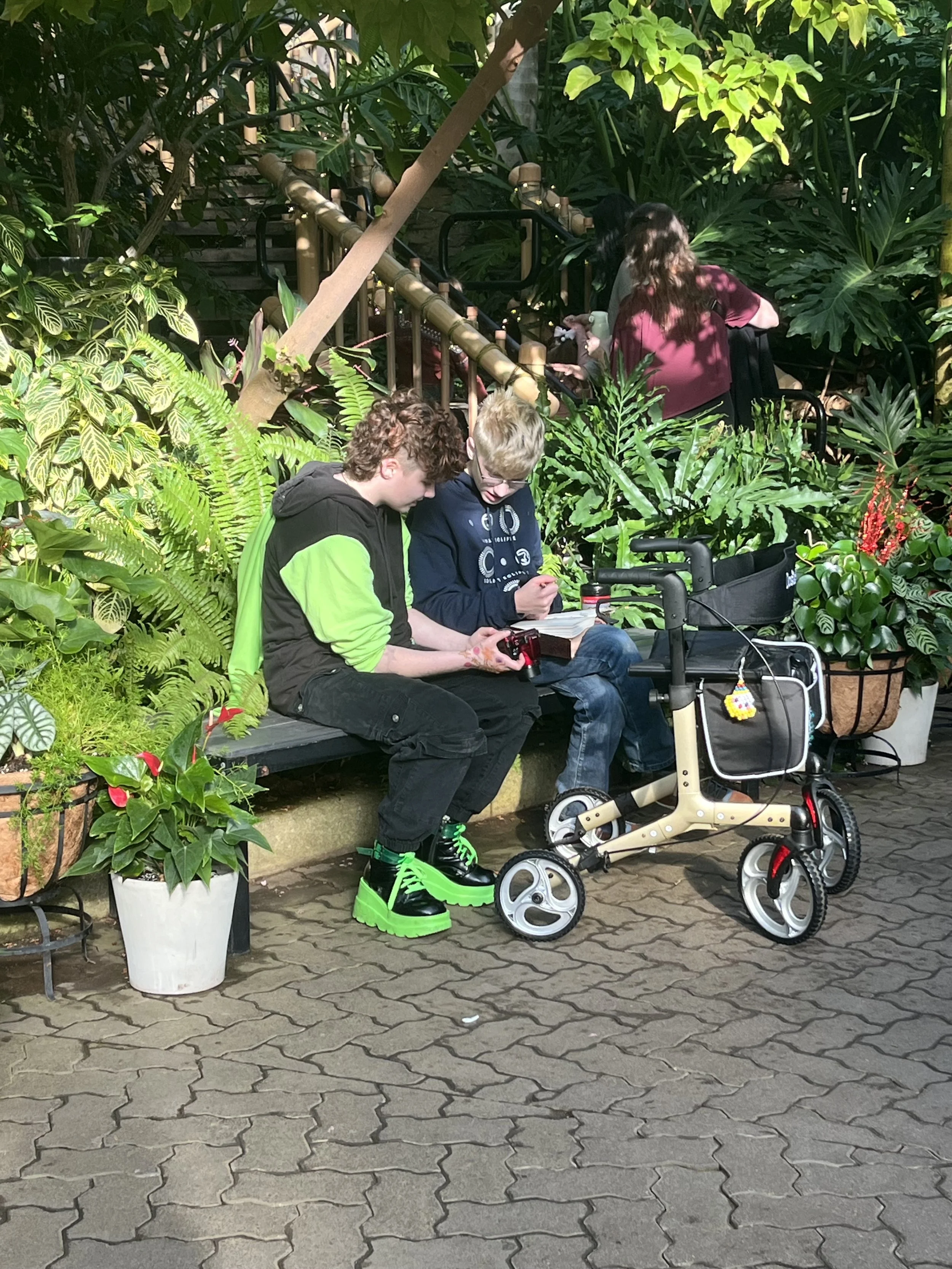 Two young men sitting on a park bench surrounded by lush green plants, one wearing black and green shoes and the other wearing glasses and a blue hoodie. A beige walker with a small yellow plush toy is beside them. In the background, a woman with long dark hair, wearing a maroon t-shirt, is walking up a staircase. The area has brick paving and plenty of greenery.