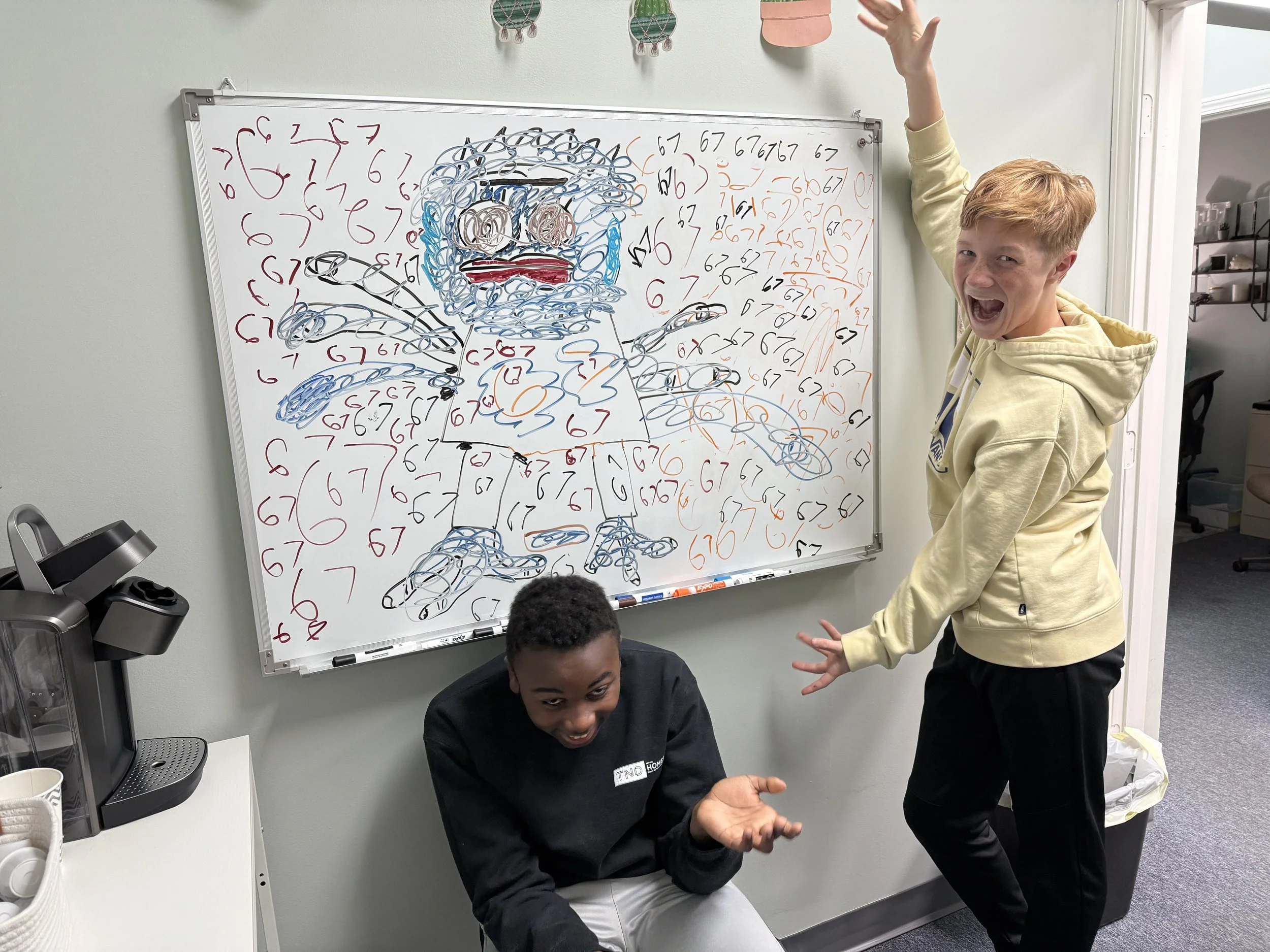 Two children, one sitting on the floor and one standing, posing playfully in front of a whiteboard with a colorful scribble drawing of a face made with numbered lines. The standing boy has red hair, and the sitting boy has black hair.