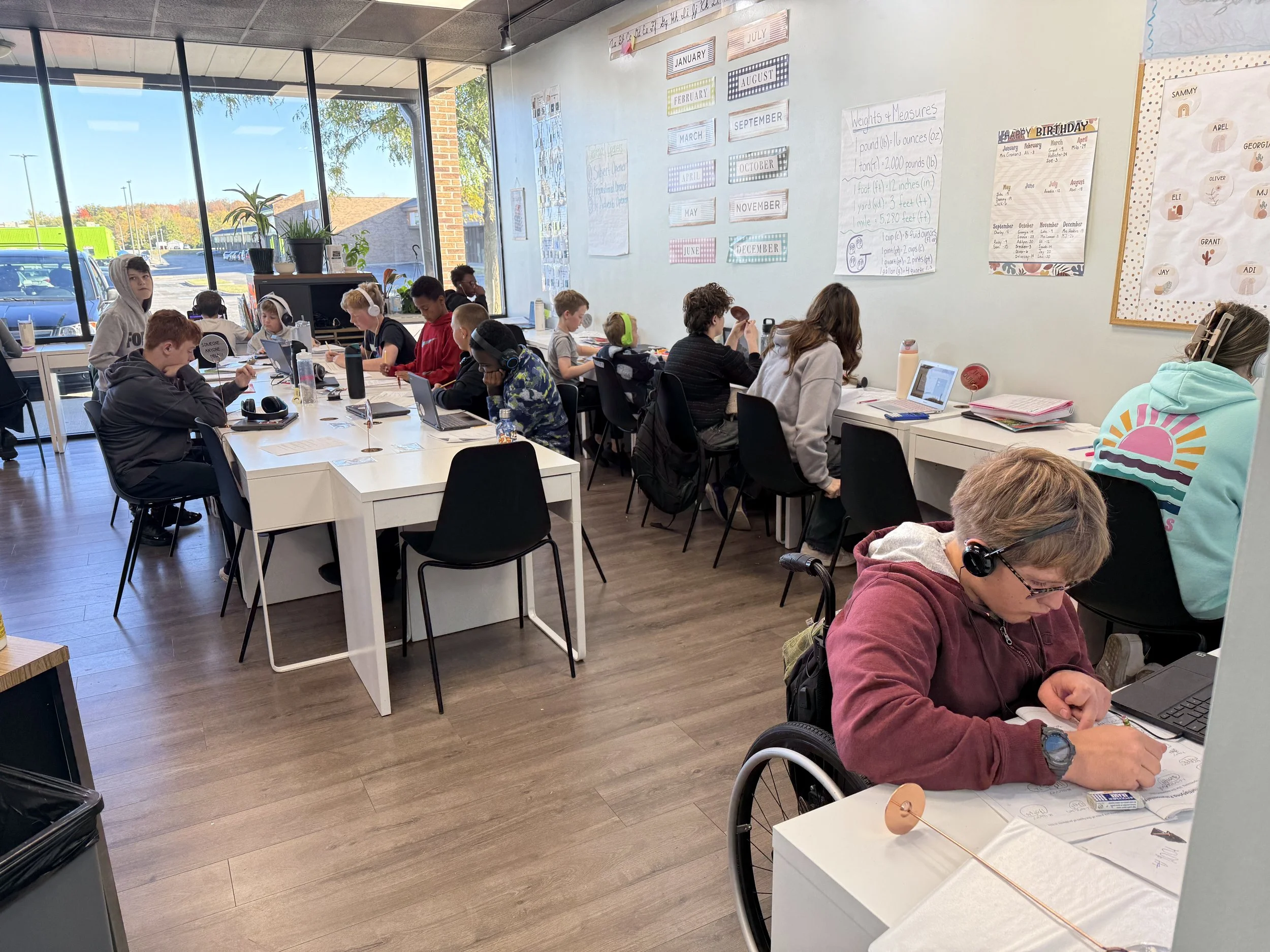 A classroom filled with students working on laptops and writing at desks, with large windows showing a parking lot and trees outside, and educational posters and calendar on the walls.