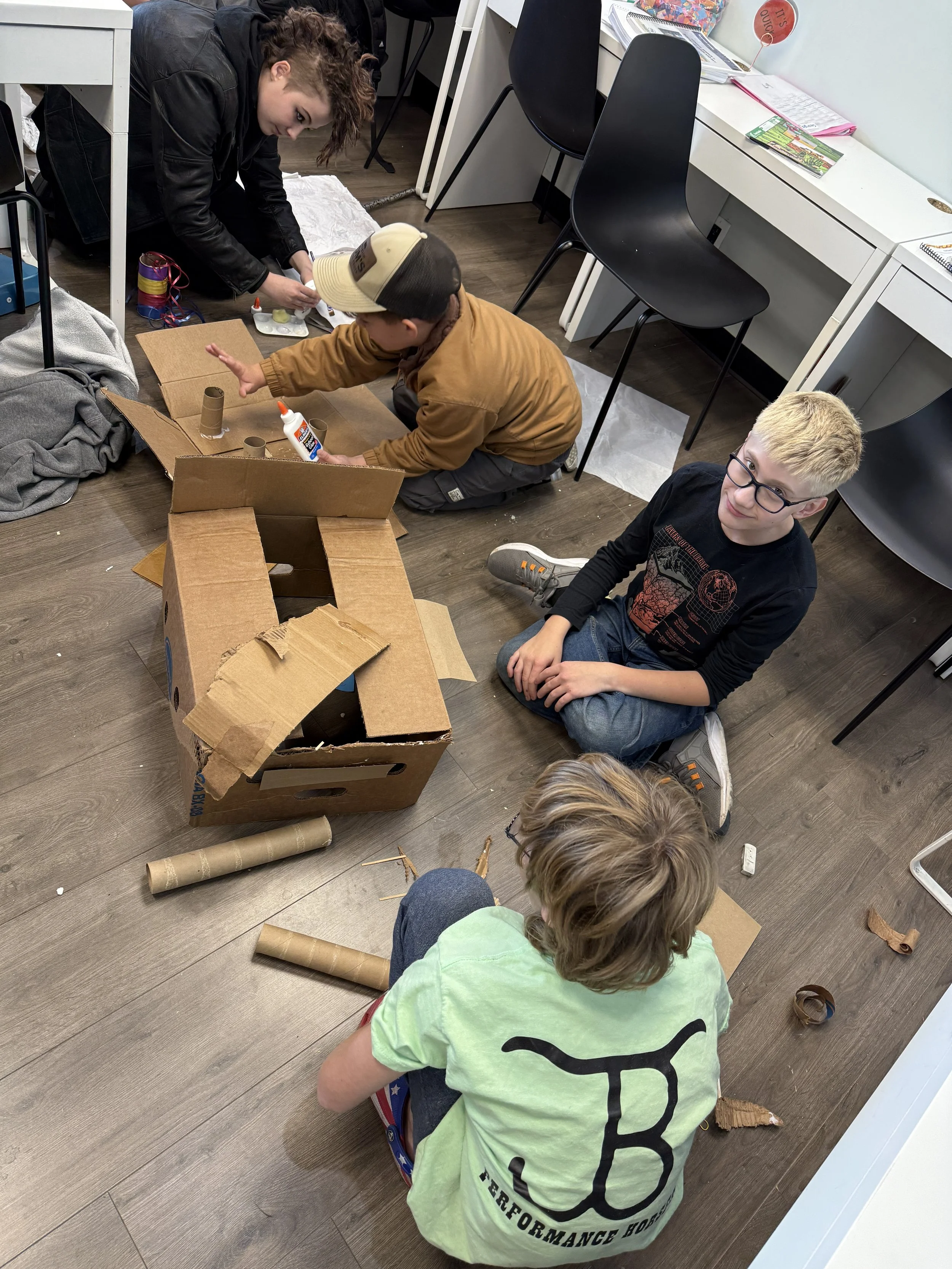 Teenagers and children making crafts with cardboard tubes and glue on a wooden floor in a classroom or craft room.