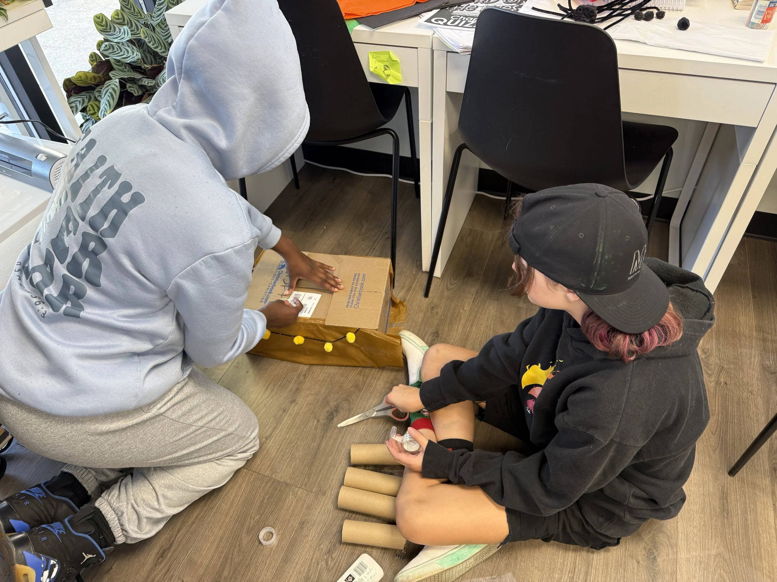Two children and an adult working on arts and crafts at a table in a room with black chairs, papers, and supplies. The children are sitting on the floor with cardboard tubes and tape, while the adult is leaning over with a box.