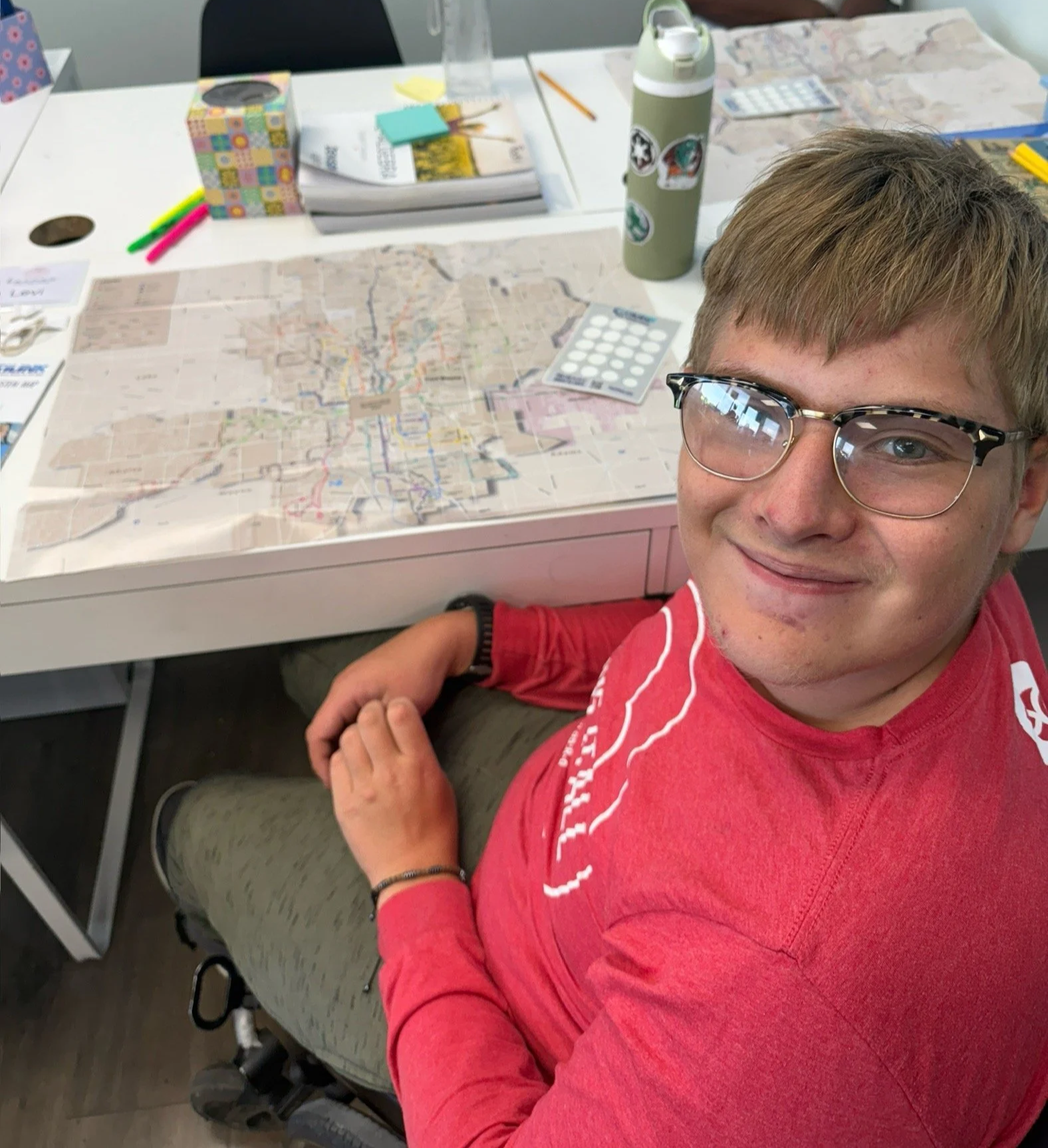 A young man with glasses and light brown hair smiling at the camera, sitting at a table with maps, pens, markers, and various office supplies. He is wearing a red shirt and beige shorts.