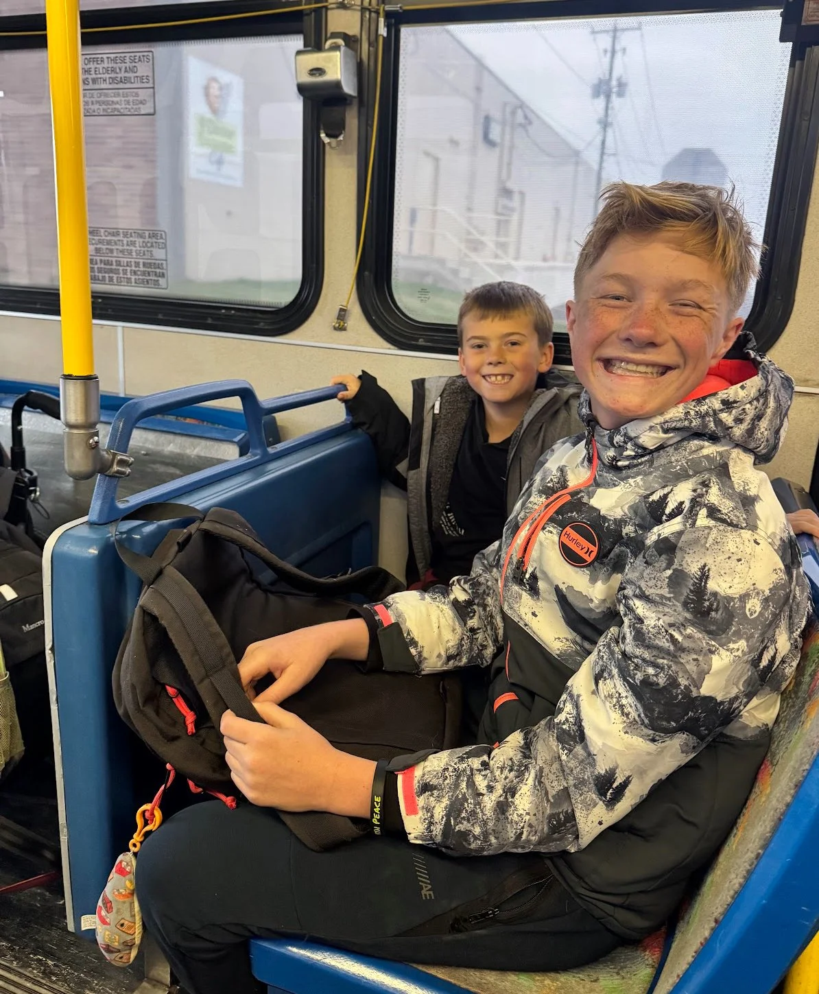 Two smiling boys sitting on a bus, one is in the foreground wearing a patterned jacket and the other behind wearing a jacket with a backpack, with buses and power lines visible outside the window.
