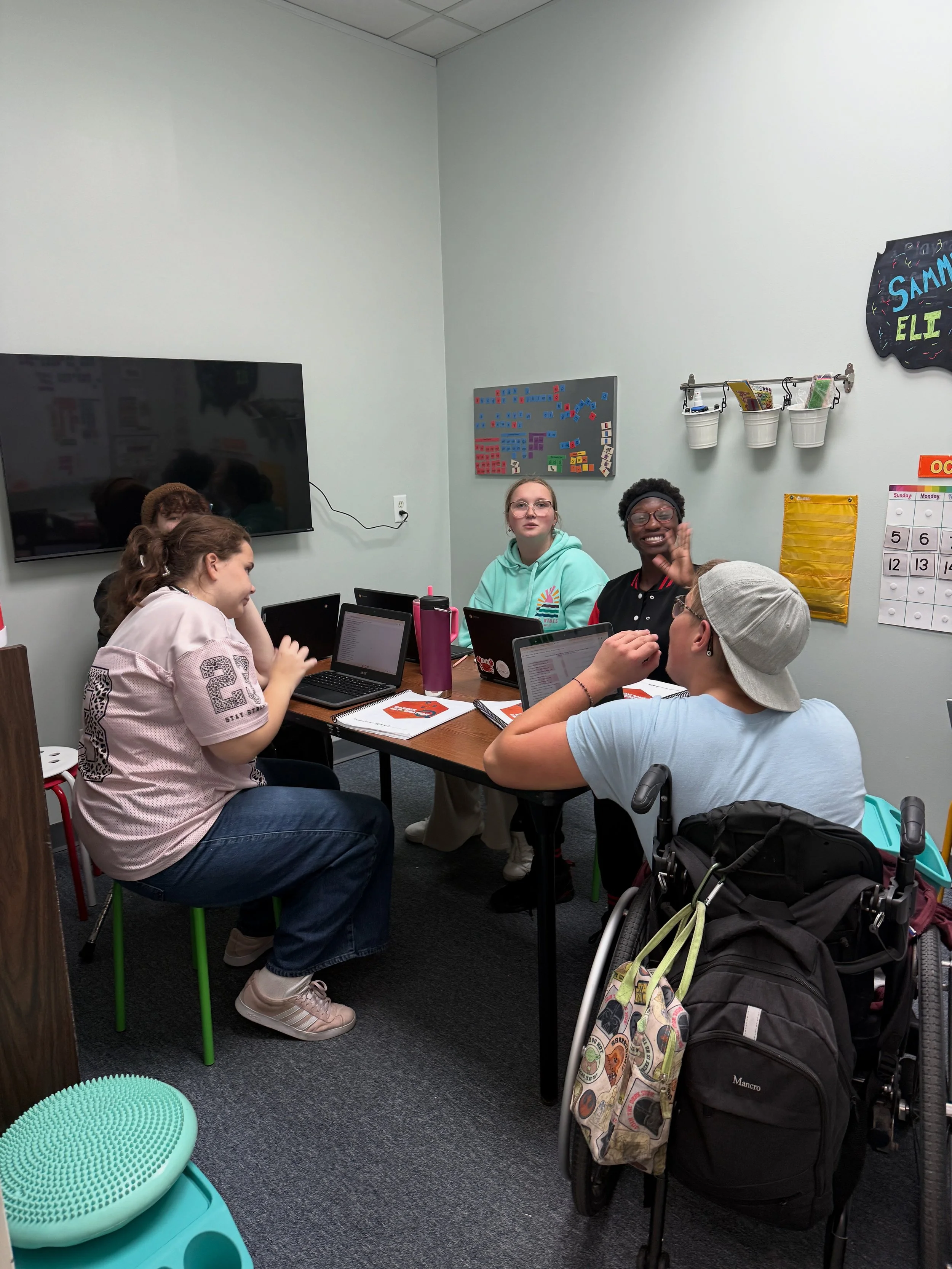 A group of young people sitting around a table in a classroom or meeting room, engaged in discussion or working on laptops, with a wheelchair nearby and colorful decorations and organizational posters on the wall.